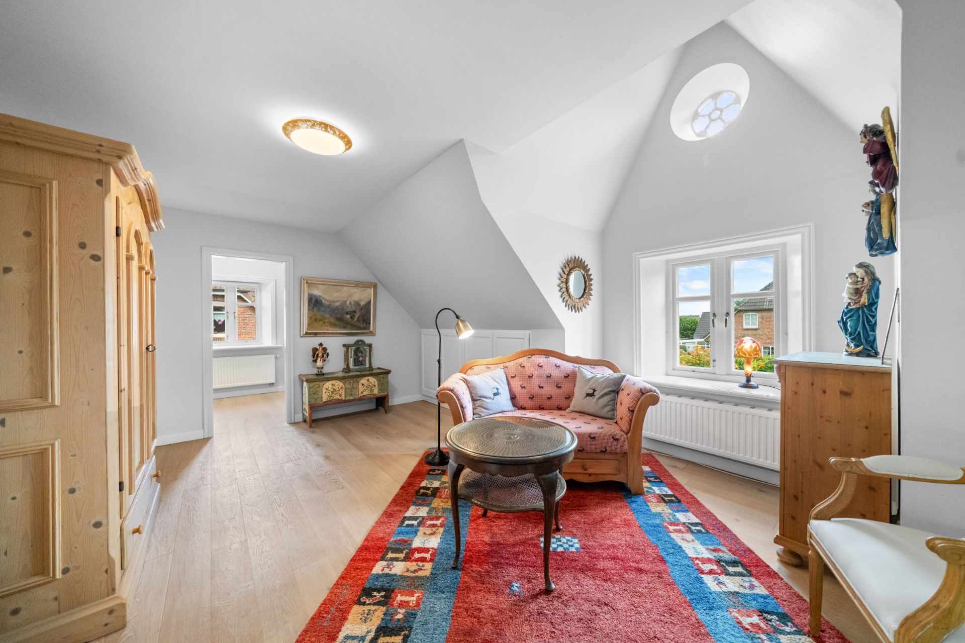 Helles Wohnzimmer mit Holzschrank, Sofa, Tisch und buntem Teppich unter schräger Decke.