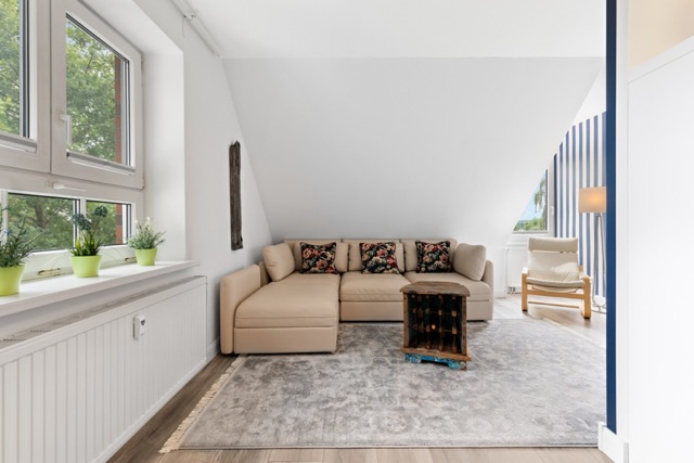 Helles Wohnzimmer mit Ecksofa, Teppich, Couchtisch und Pflanzen auf der Fensterbank.