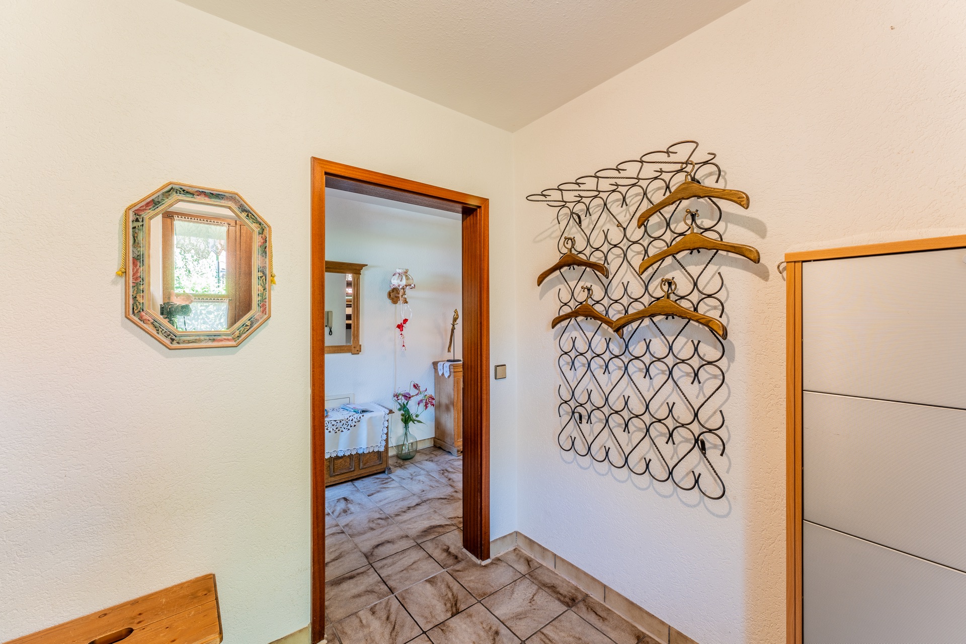 Flur mit Wandgarderobe, Kleiderbügeln, Spiegel und Blick in ein weiteres Zimmer.