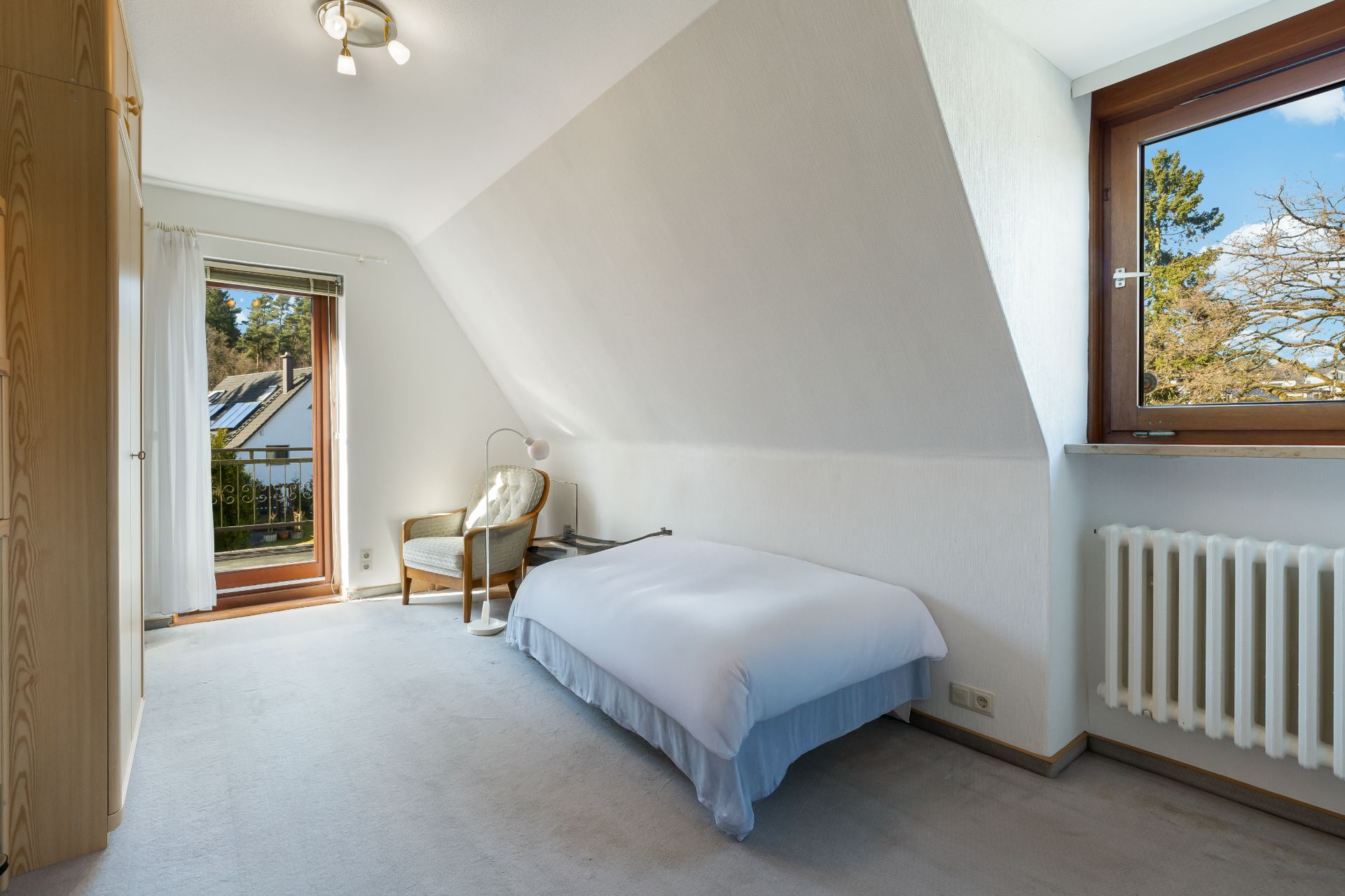 Helles Schlafzimmer mit Einzelbett, Stuhl, Kleiderschrank, Fenster und Tür zum Balkon.