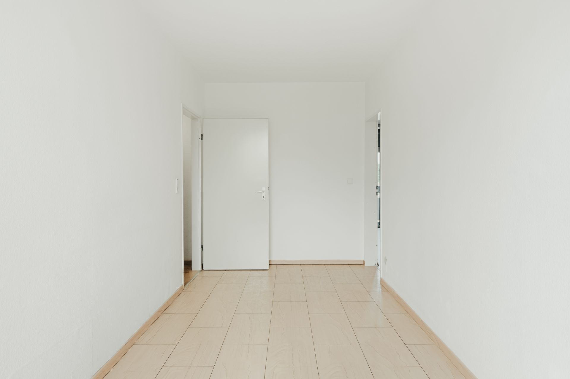 Leerer, weiß gestrichener Raum mit Fliesenboden und einer offenen Tür an der Rückwand.