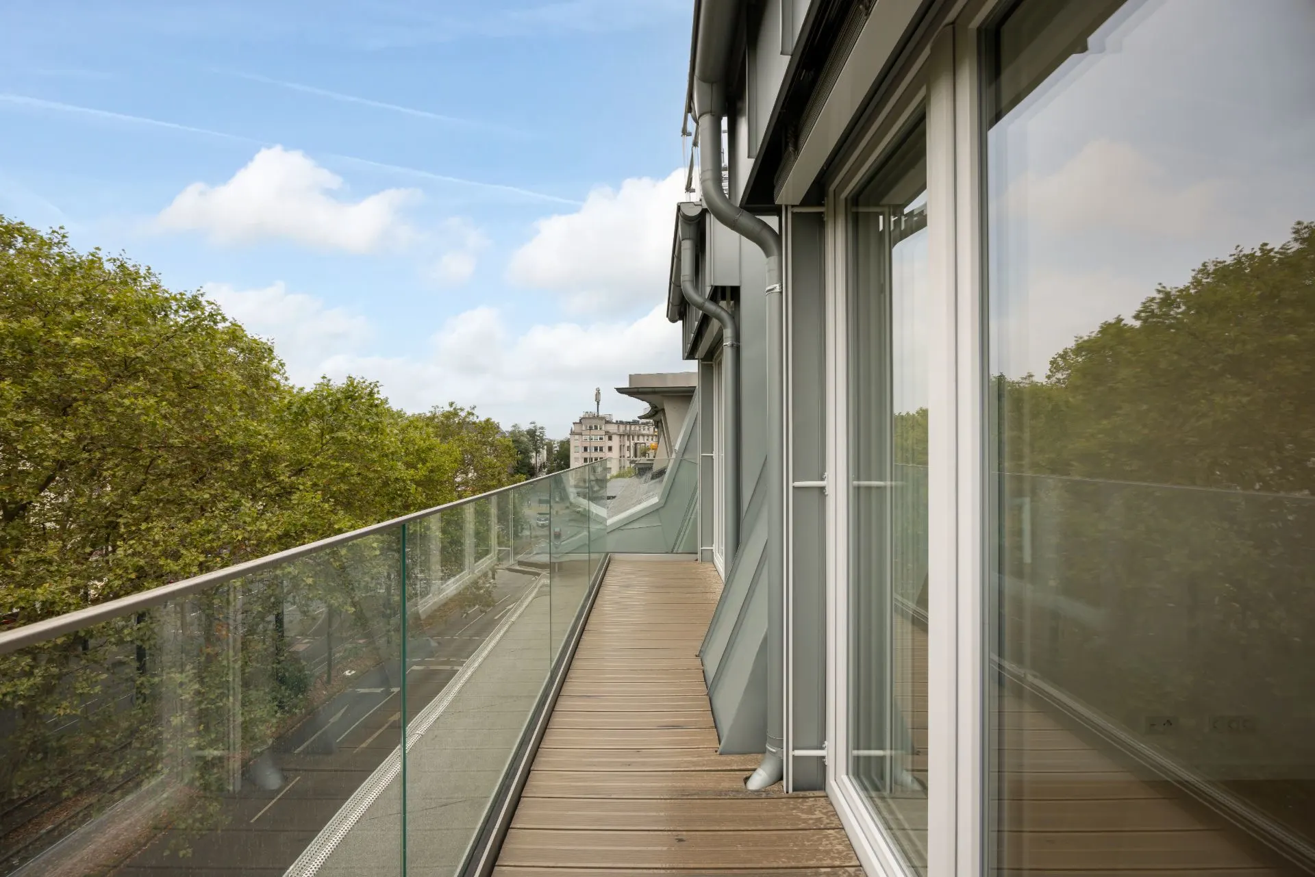 Langer Balkon mit Glasgeländer an einem Wohngebäude, Blick auf Bäume und Straße.