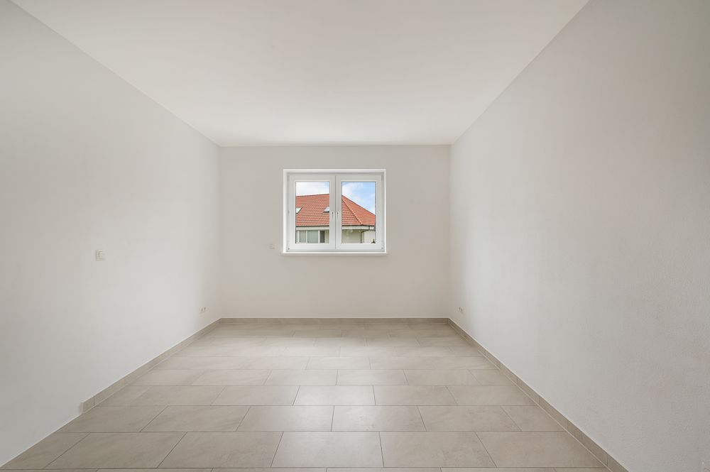 Leerer, weiß gestrichener Raum mit Fliesenboden und Fenster mit Blick auf ein Hausdach.