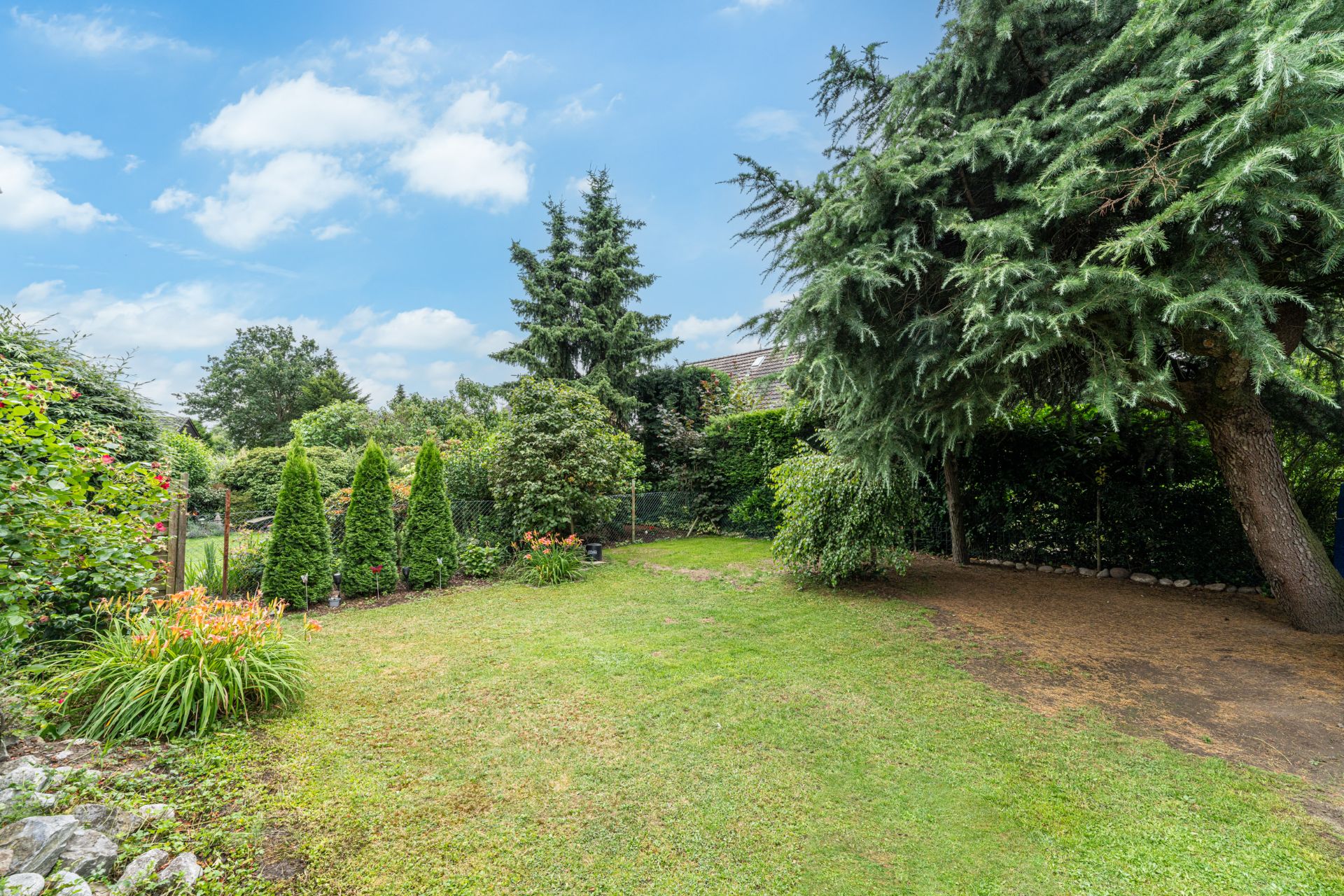 Grüner Garten mit Rasen, Blumen, Sträuchern und großen Bäumen unter blauem Himmel.