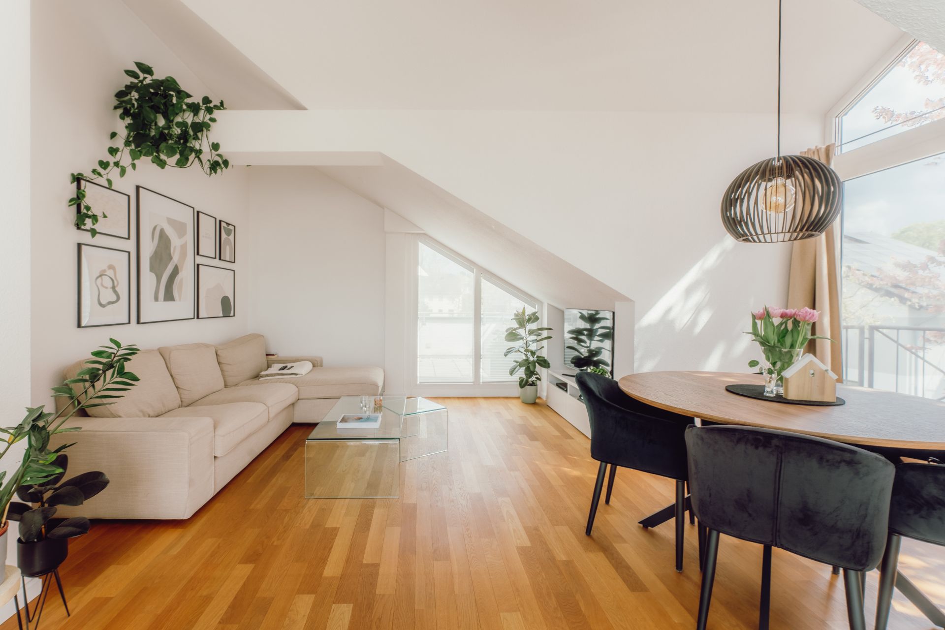 Helles Wohnzimmer mit Sofa, Esstisch, Pflanzen und großem Fenster unter Dachschräge.