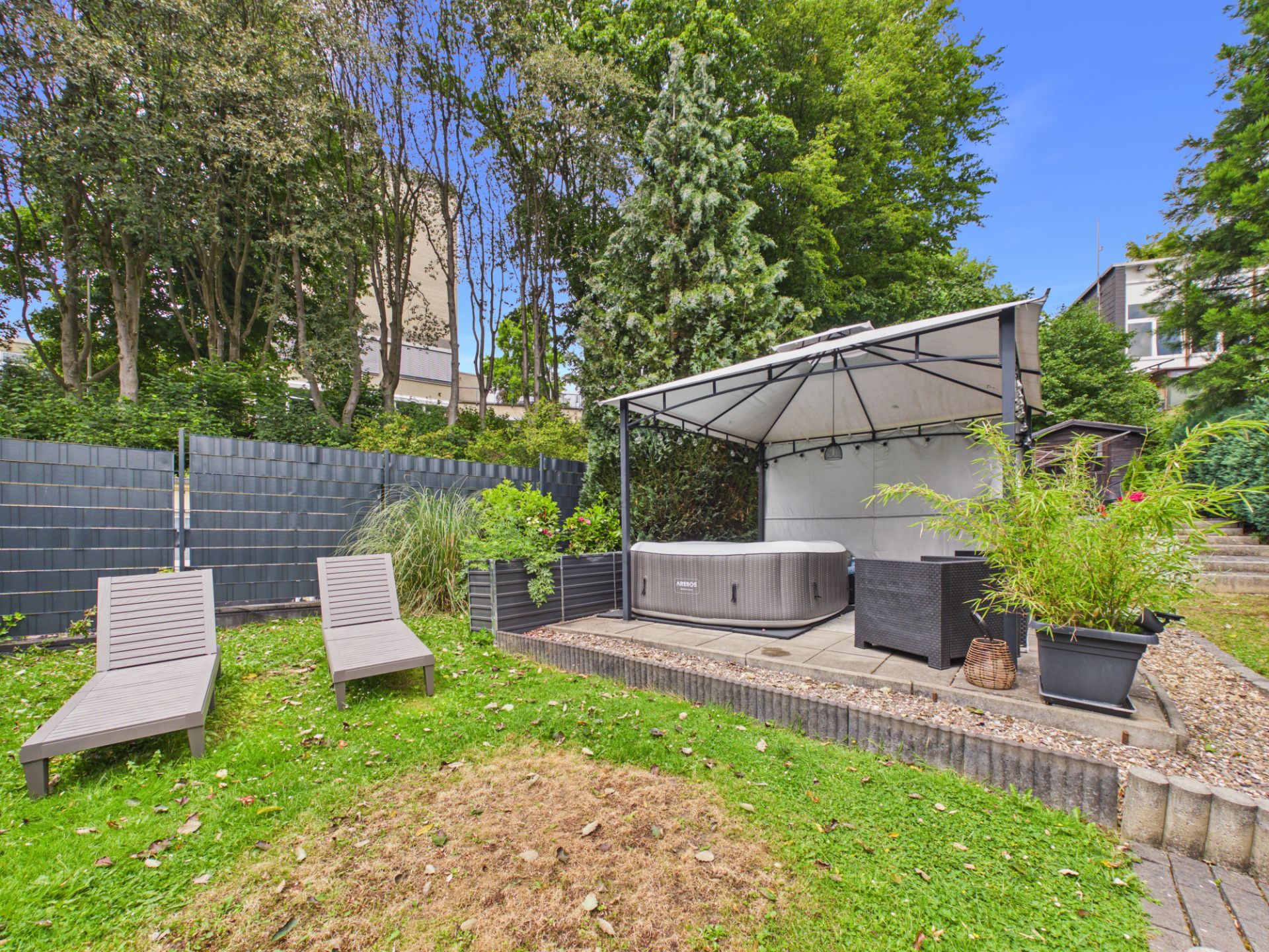 Garten mit zwei Liegestühlen und überdachtem Whirlpool auf einer Terrasse, umgeben von Pflanzen.