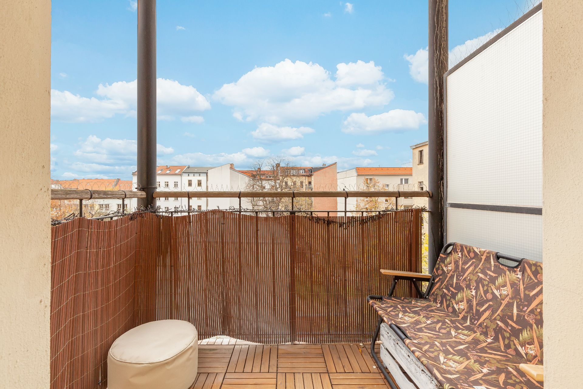 Kleiner Balkon mit Holzlattenboden, Sitzbank mit gemustertem Bezug und Hocker, Ausblick auf Wohnhäuser.