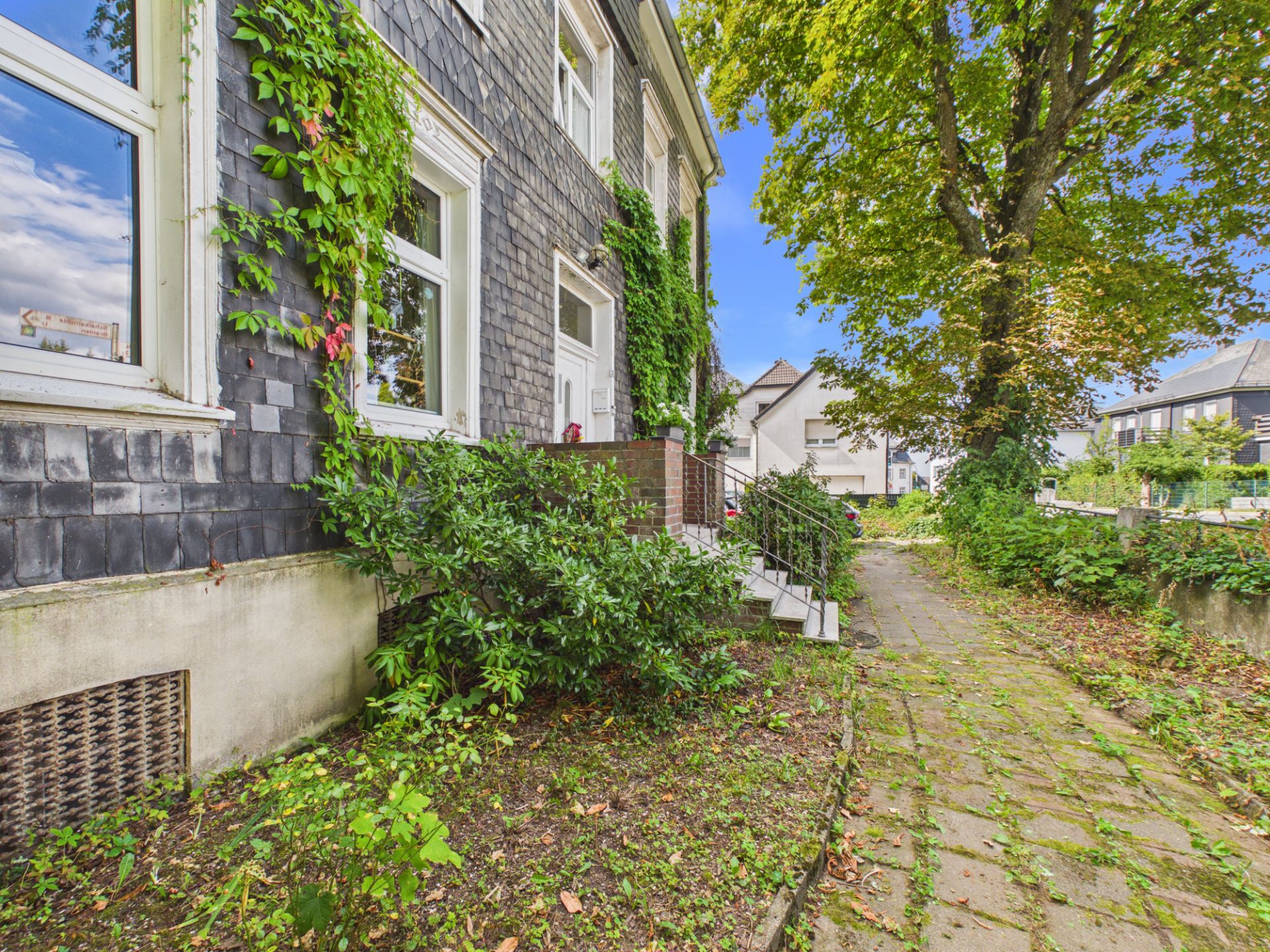 Ein Weg führt an einem Haus mit grauer Fassade und grünen Pflanzen entlang.