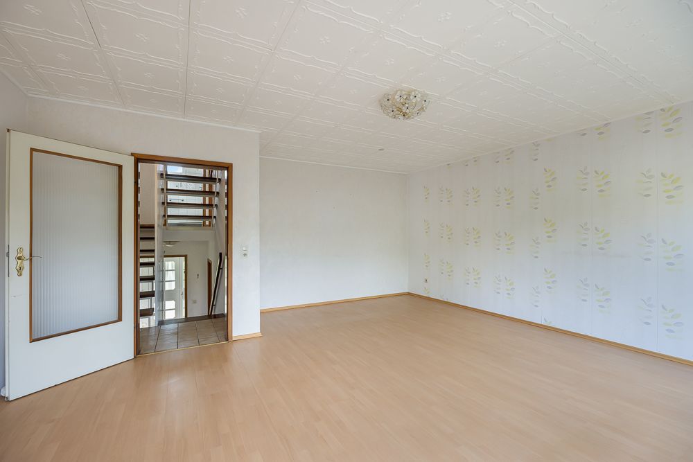 Leeres Zimmer mit hellem Holzboden, gemusterter Tapete und Blick auf Treppenhaus.