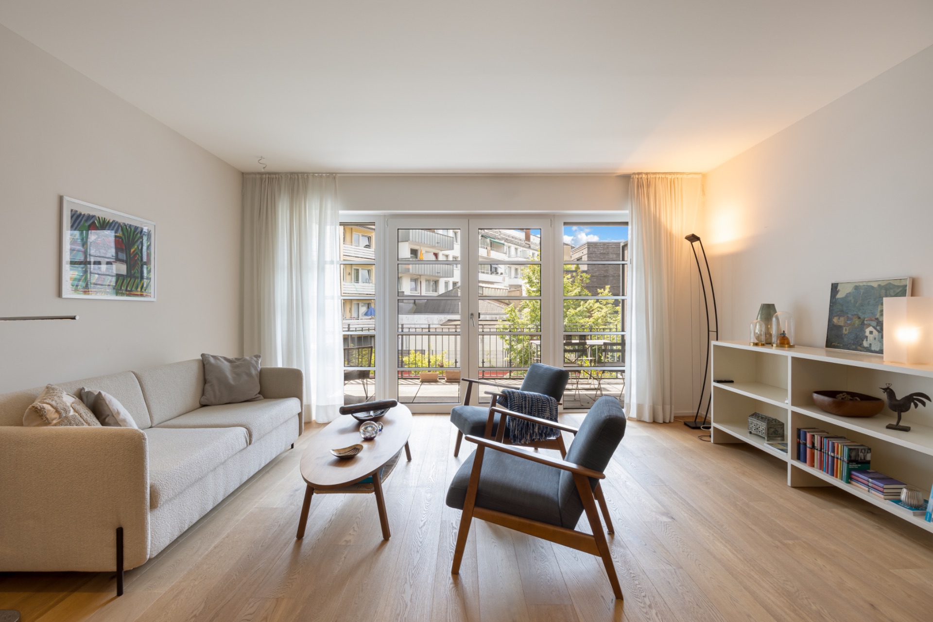 Helles Wohnzimmer mit Sofa, zwei Sesseln, Couchtisch und Regal, große Fenster mit Blick auf Balkon.