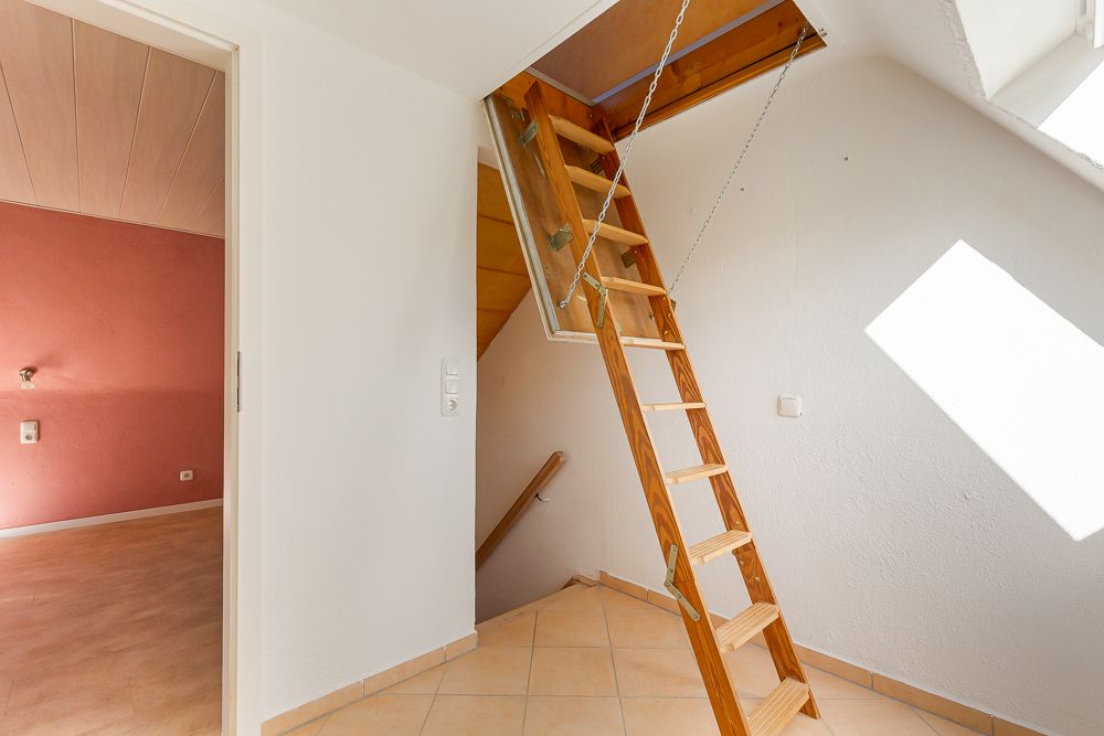 Holzklappleiter führt zu einer Dachbodenluke in einem hellen Flur mit Fliesenboden.