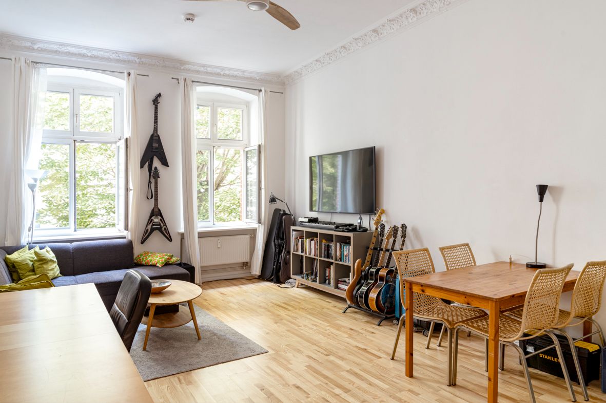 Wohnzimmer mit Sofa, Esstisch, Fernseher, Gitarren und großen Fenstern mit weißen Vorhängen.