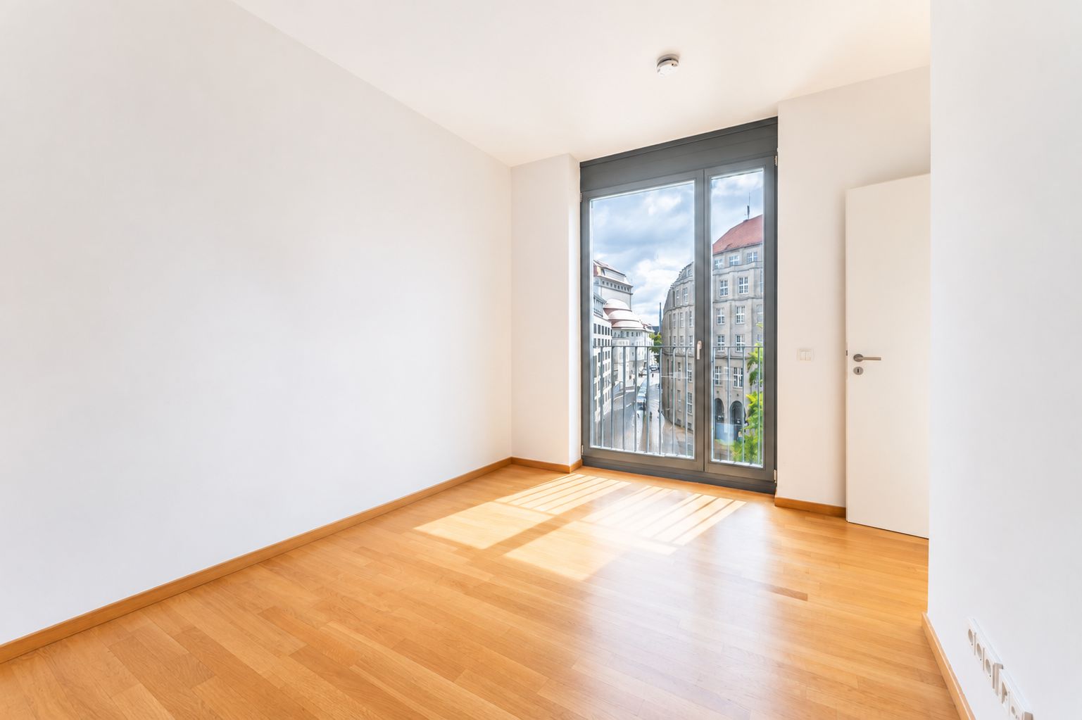 Heller, leerer Raum mit Holzboden, weißer Wand und großem Fenster mit Stadtblick.