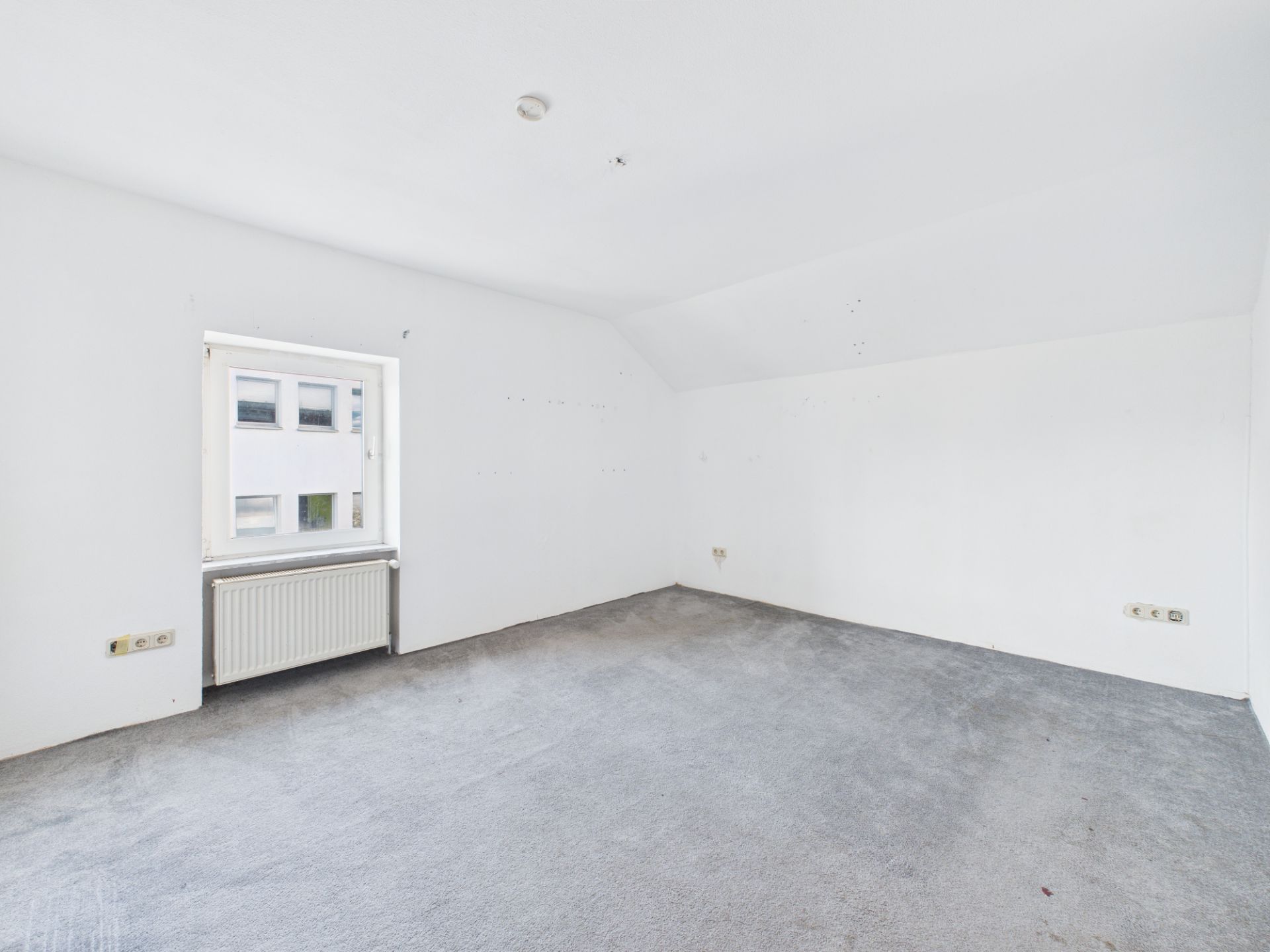 Leeres Zimmer mit weiß gestrichenen Wänden, grauem Teppich und einem Fenster mit Heizkörper.