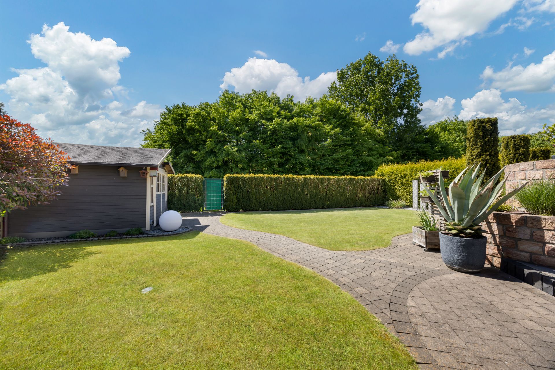 Gepflegter Garten mit Rasen, Gartenhaus und gepflastertem Weg bei sonnigem Wetter.