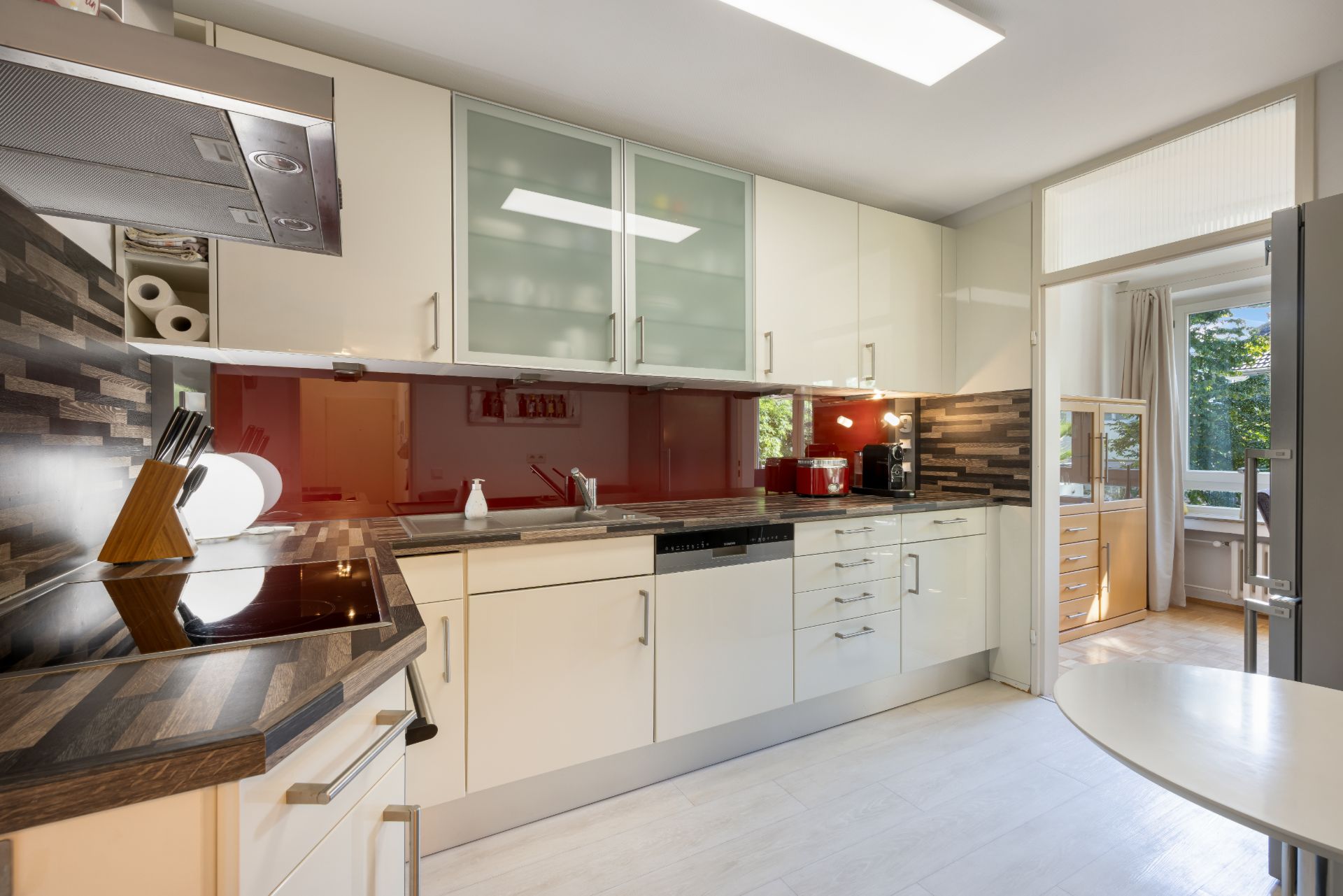 Moderne Küche mit weißen Schränken, roter Glasrückwand und Holzakzent, Blick ins helle Nebenzimmer.