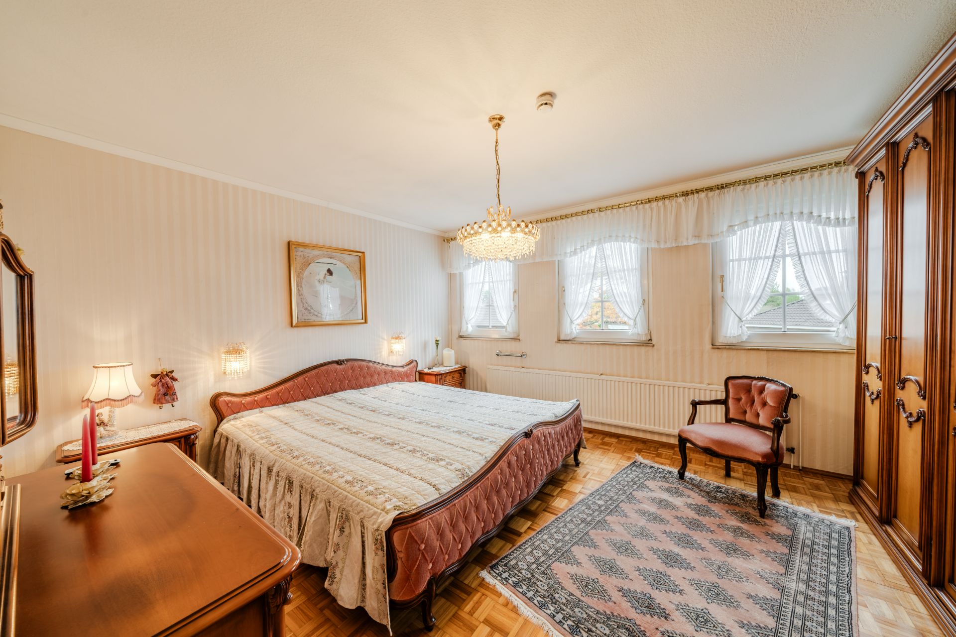 Helles Schlafzimmer mit großem Bett, Kronleuchter, Teppich, Holzschrank und drei Fenstern mit Gardinen.