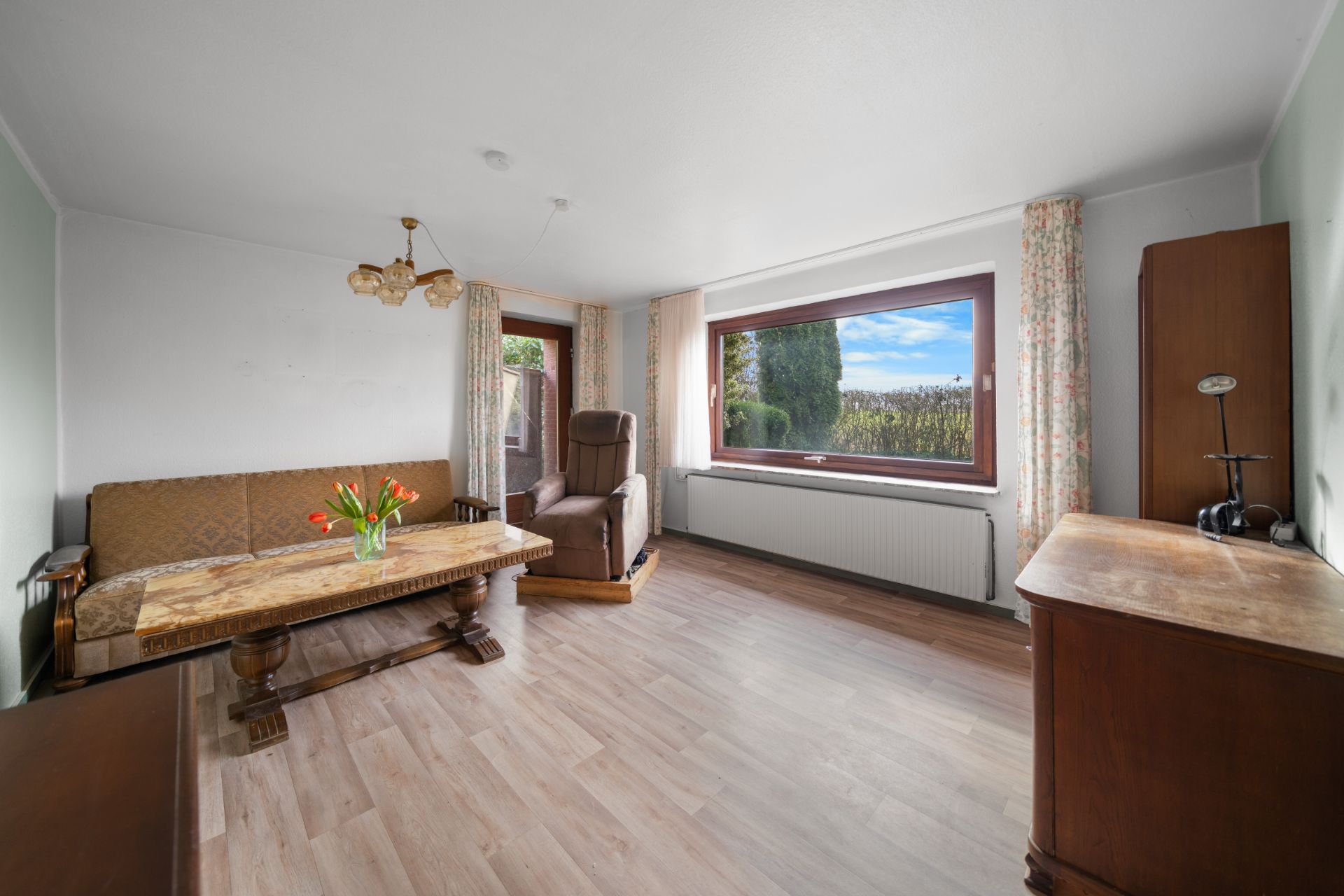 Helles Wohnzimmer mit Sofa, Sessel, Holztisch, Kommode und großem Fenster mit Blick ins Grüne.