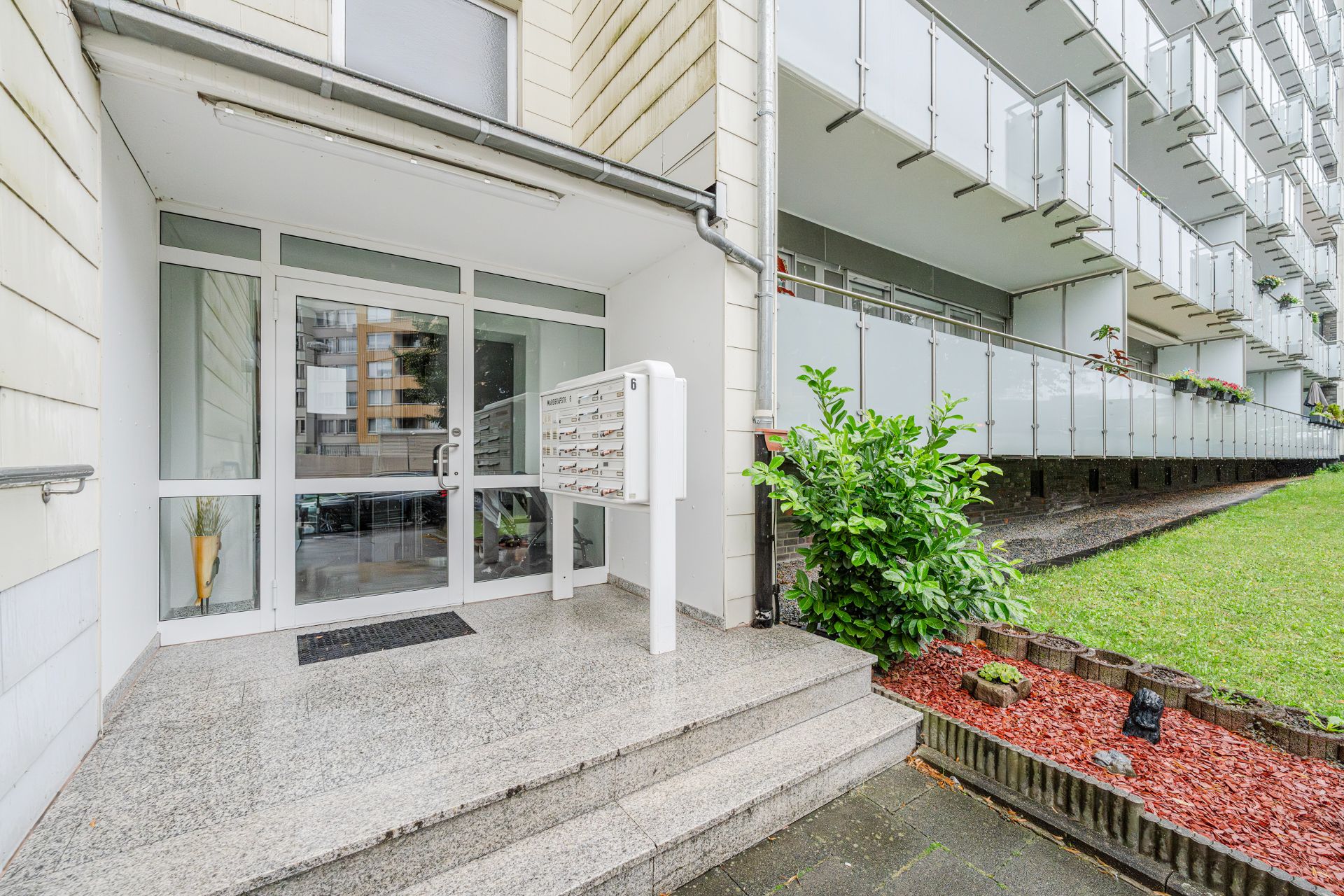Hauseingang mit Briefkästen, Treppe, Glasfront und angrenzendem Gartenbereich.