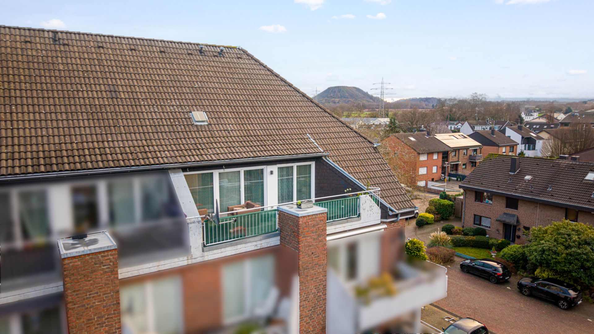 Dachgeschosswohnung mit Balkon in einem Wohnhaus, umliegende Häuser und Autos sichtbar.