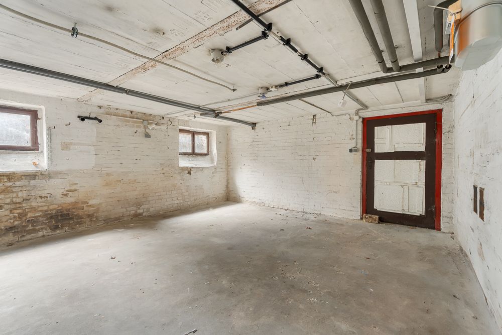 Leerer Kellerraum mit weißen Wänden, zwei kleinen Fenstern und sichtbaren Rohrleitungen an der Decke.