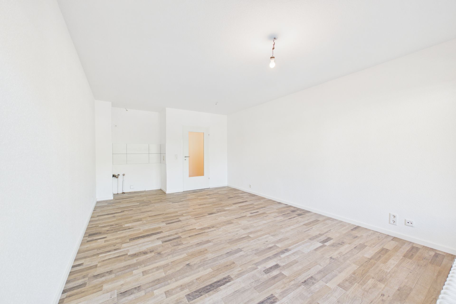 Heller, leerer Raum mit Holzboden, weißen Wänden und einer Tür.