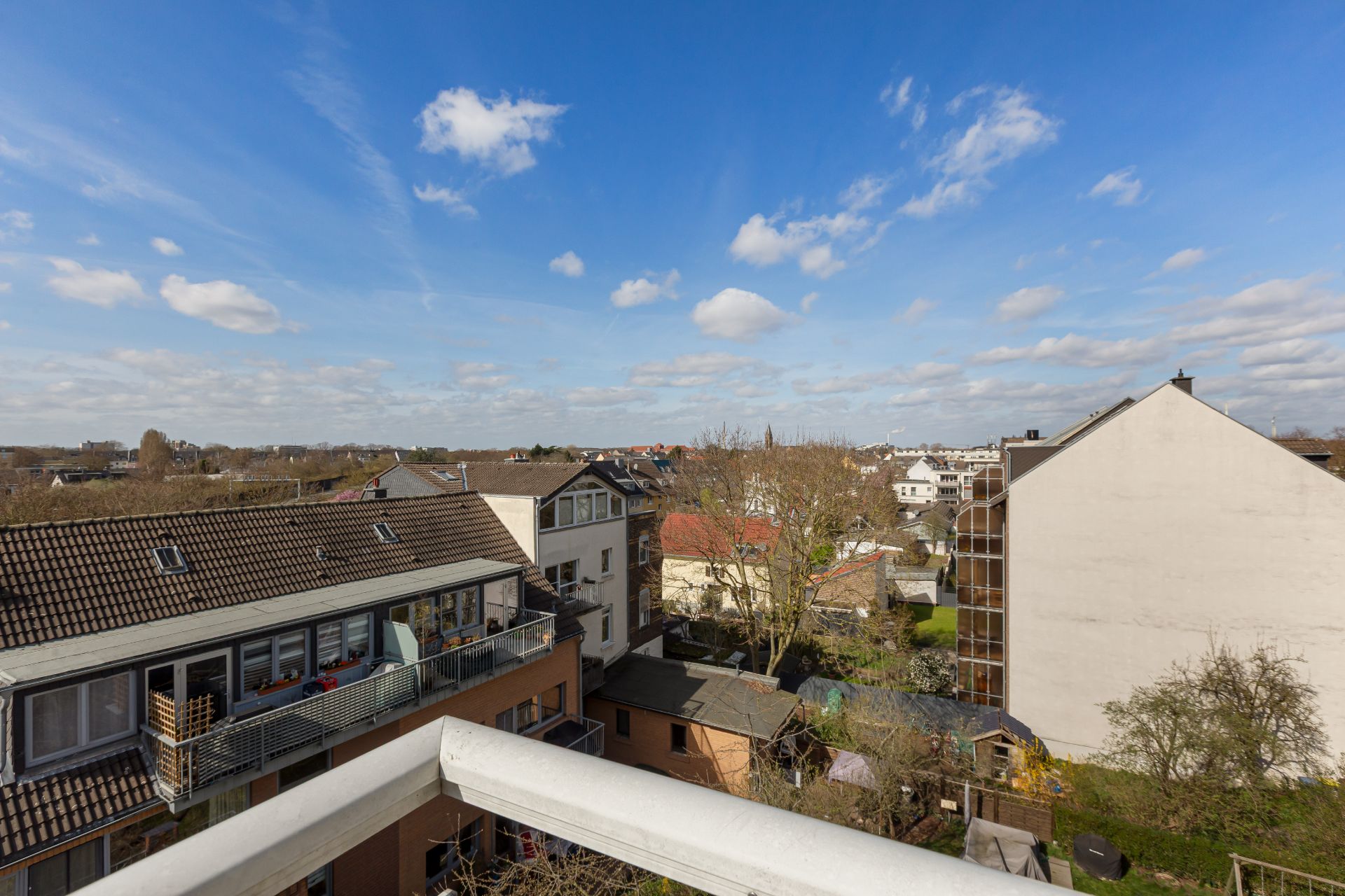 Blick vom Balkon auf Nachbarhäuser und Gärten unter blauem Himmel mit Wolken.