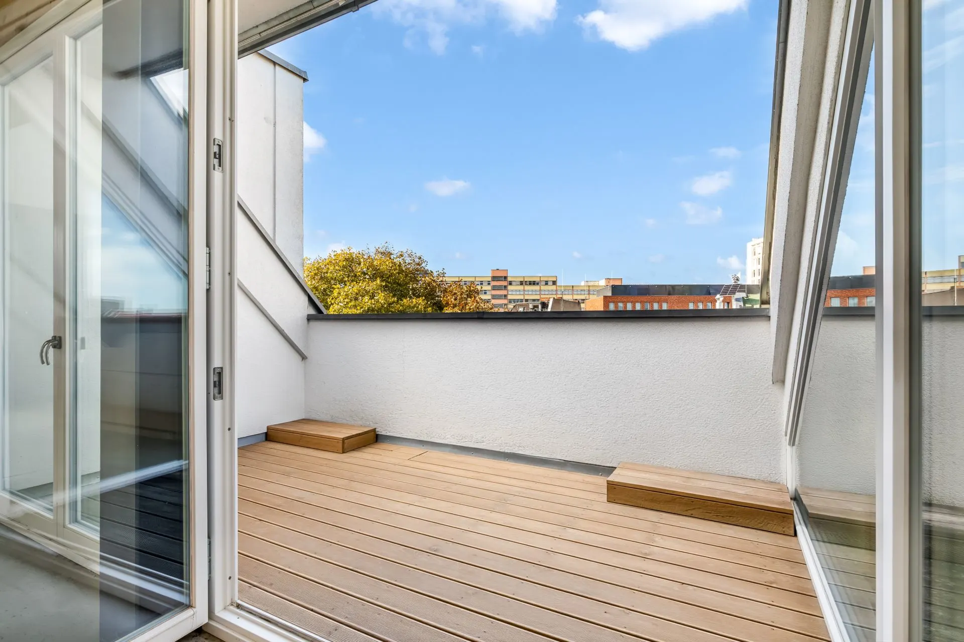 Balkon mit Holzfußboden und zwei Stufen, umgeben von weißer Mauer, Blick in die Stadt.