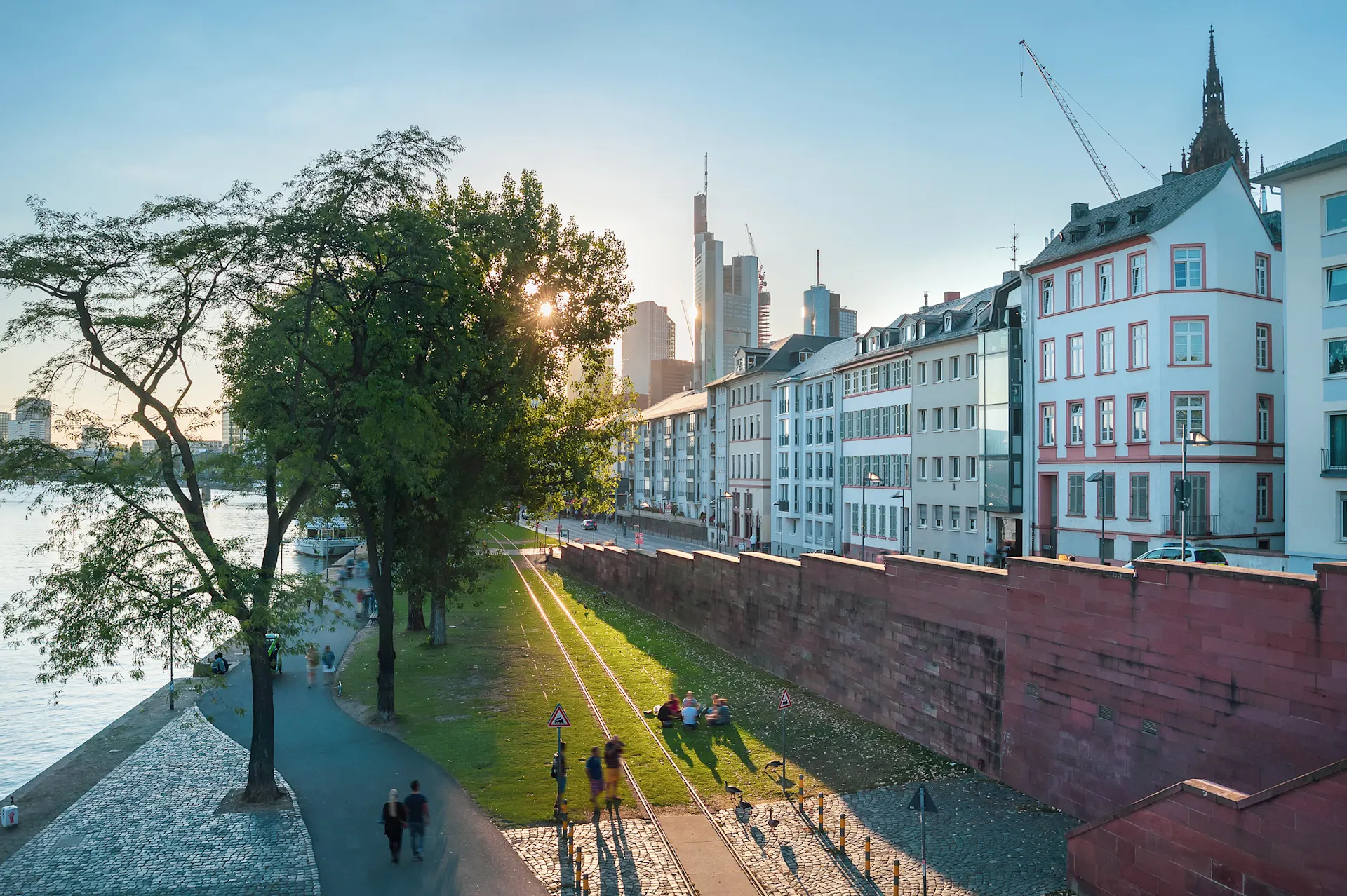 Wohnhäuser an einer Straße mit Bäumen, Park und Menschen am Flussufer bei Sonnenuntergang.