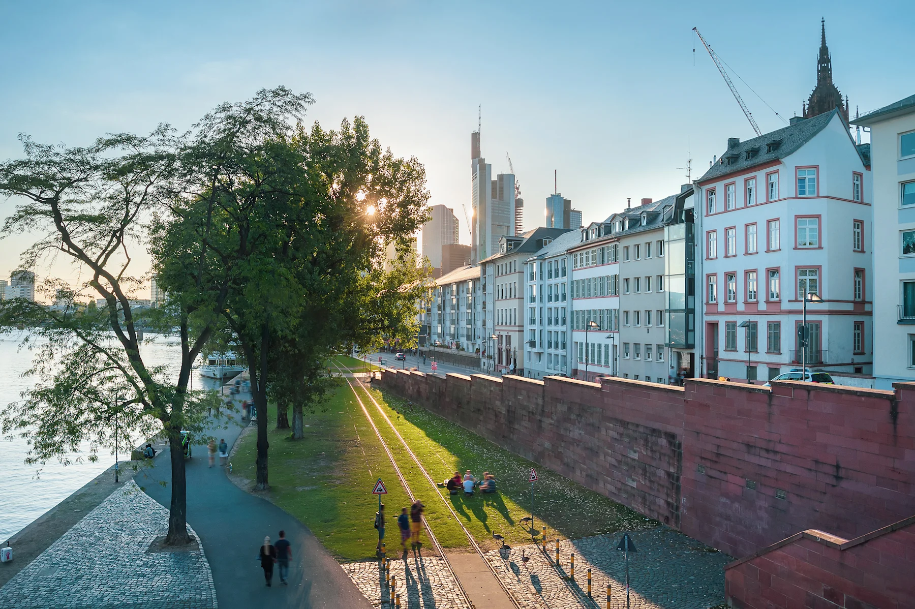 Wohnhäuser an einer Straße mit Bäumen, Park und Menschen am Flussufer bei Sonnenuntergang.