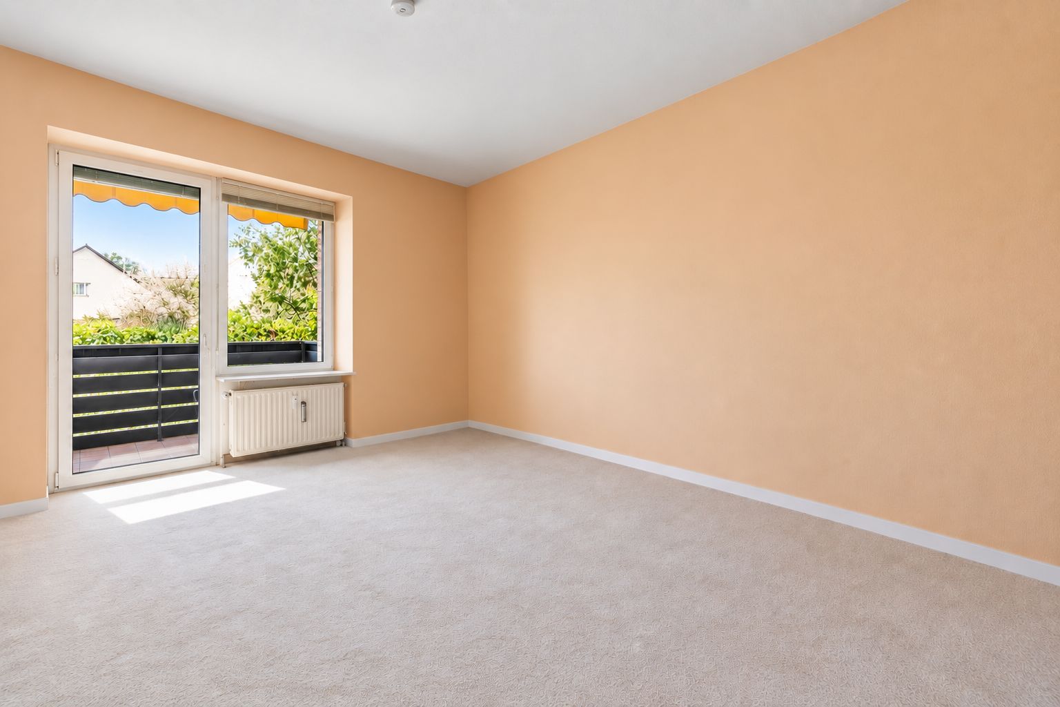 Helles, leeres Zimmer mit beigen Wänden, Teppichboden und Zugang zu einem Balkon.