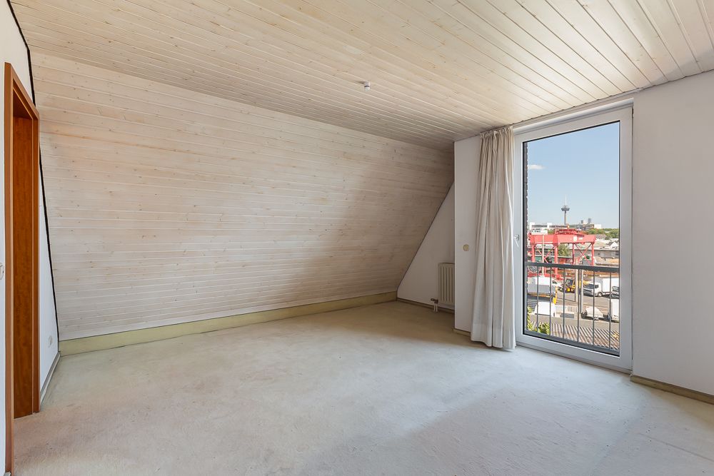 Leeres Zimmer mit schräger Holzdecke, großem Fenster und Blick nach draußen.