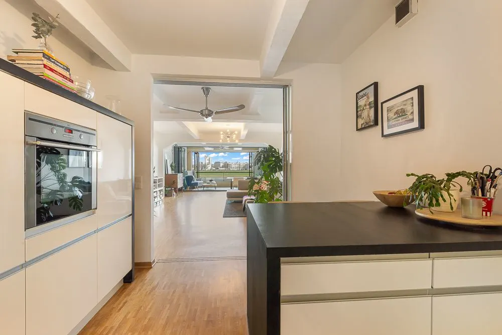 Blick von einer modernen Küche in ein helles Wohnzimmer mit Holzfußboden und großen Fenstern.