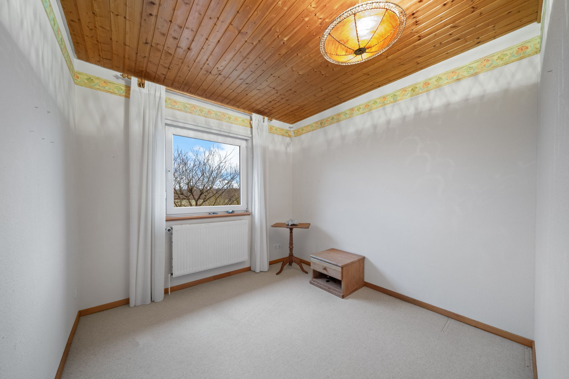 Kleines Zimmer mit Holzbalkendecke, Fenster, weißen Wänden, Vorhängen und zwei kleinen Möbelstücken.