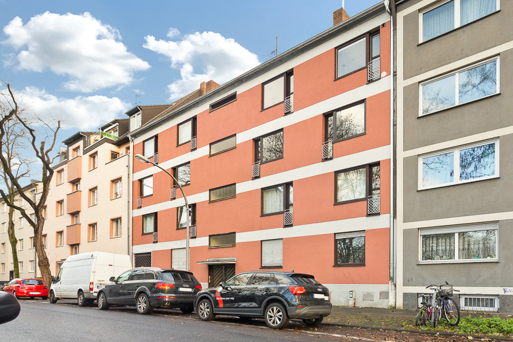 Mehrstöckiges Wohngebäude mit roten und grauen Fassaden, geparkte Autos und Fahrräder davor.