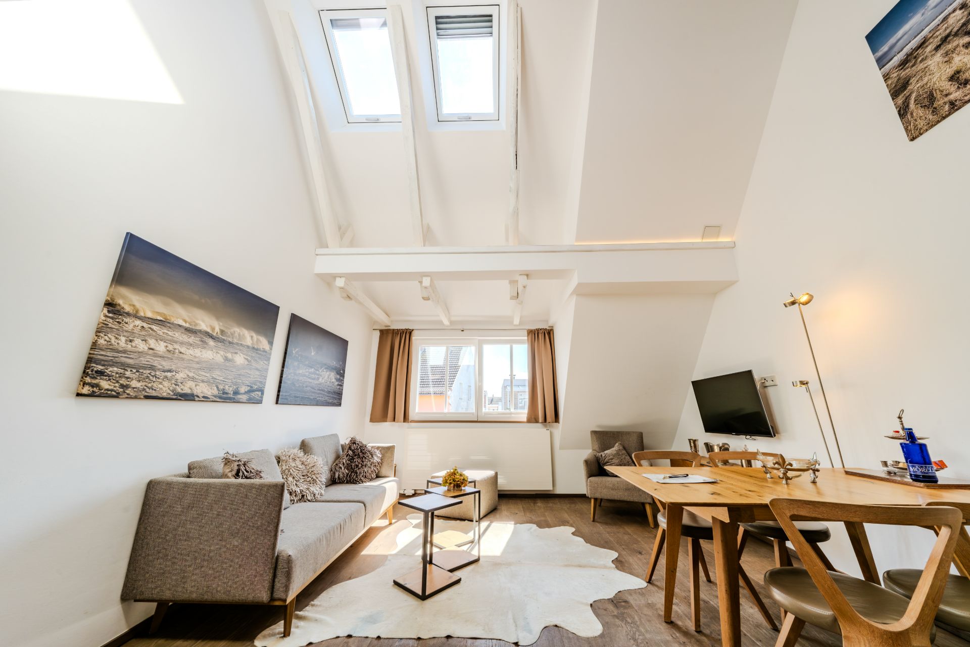 Helles Wohnzimmer mit Sofa, Esstisch, Wandbildern und Dachfenstern.