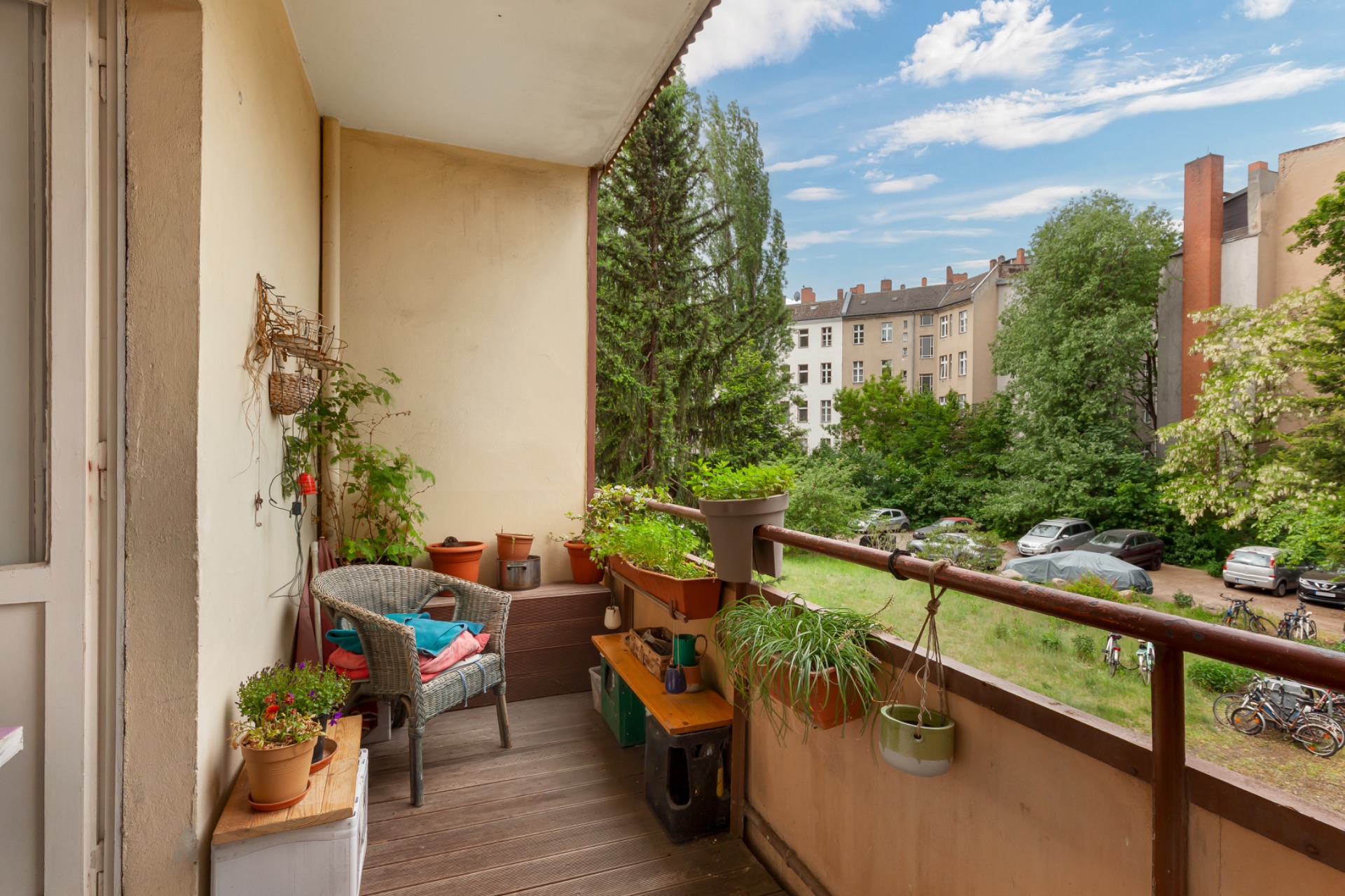 Balkon mit Pflanzen, Stuhl und Blick auf einen grünen Innenhof mit Bäumen und geparkten Autos.