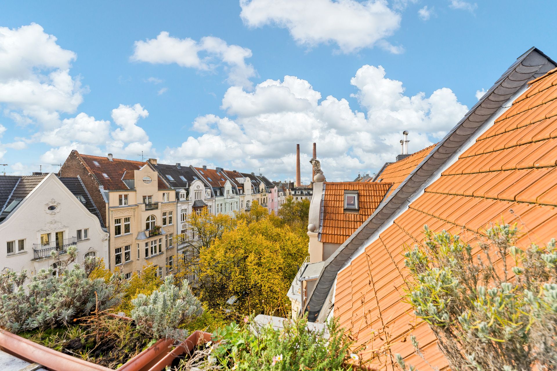 Blick von einem Balkon auf Dächer, Bäume und Wohnhäuser unter blauem Himmel mit Wolken.
