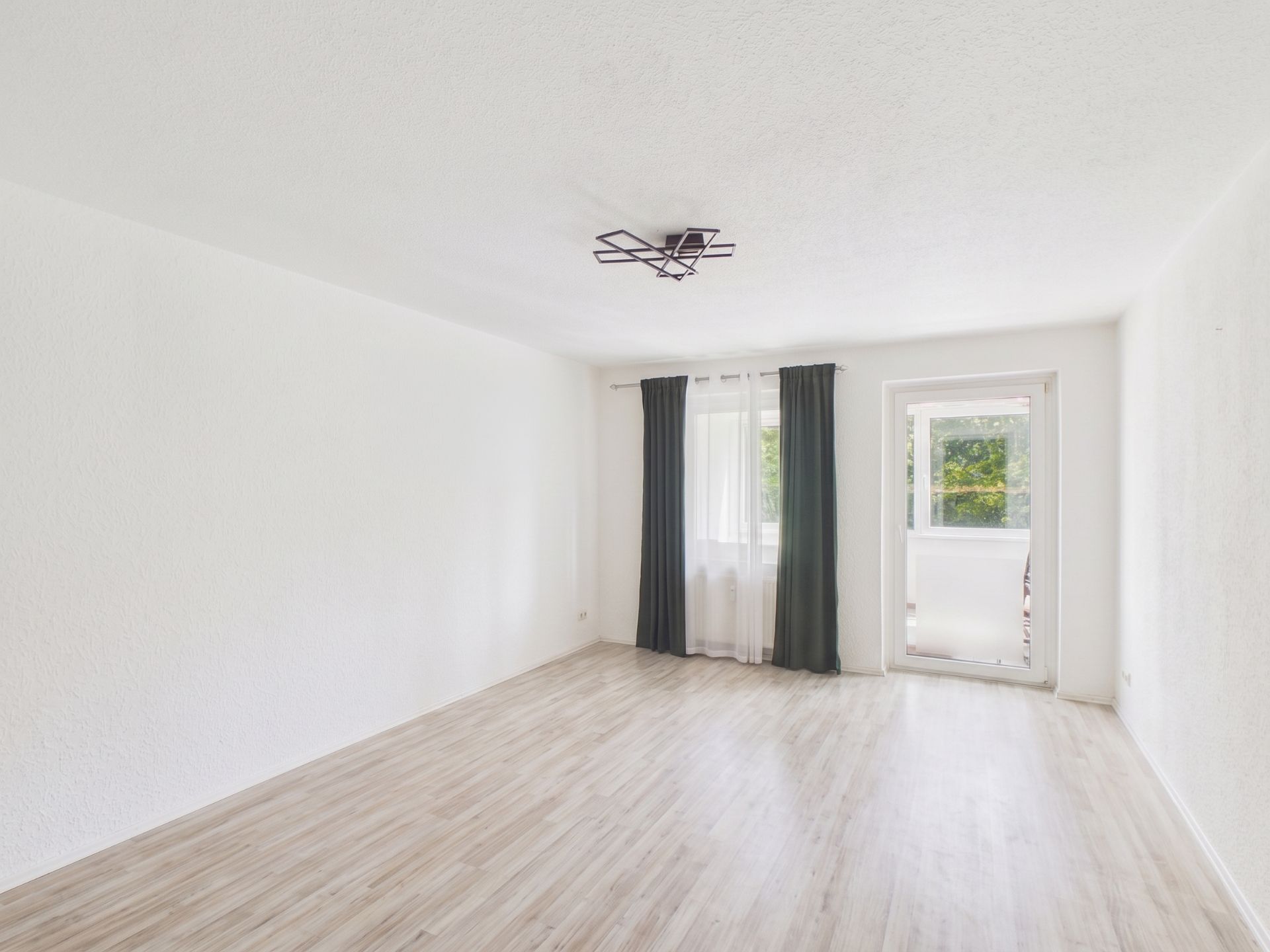 Helles, leeres Zimmer mit hellem Boden, Fenster, Vorhängen und Zugang zu einem Balkon.