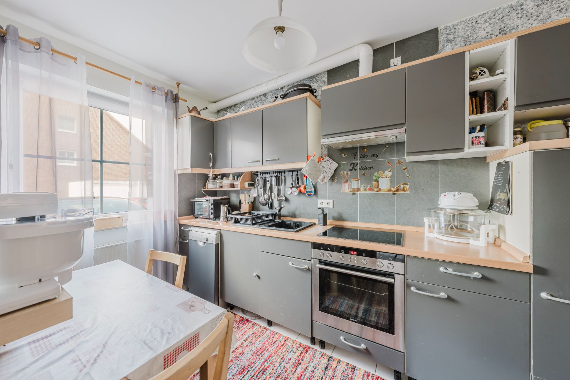 Moderne Küche mit grauen Schränken, Herd, Esstisch und großem Fenster mit Gardinen.
