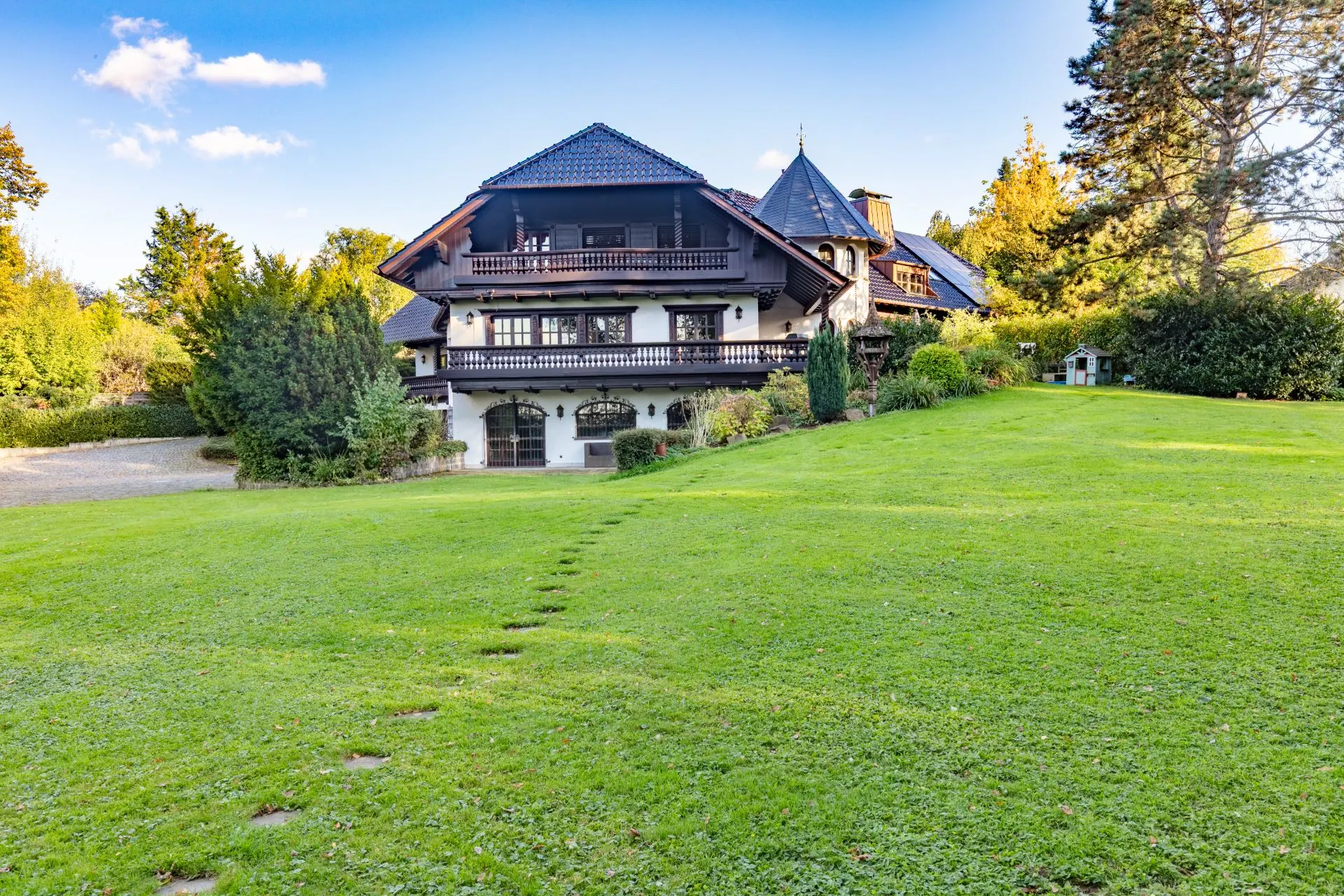 Großes Haus mit dunklem Dach, Balkon und breitem Rasen davor, umgeben von Bäumen und Büschen.