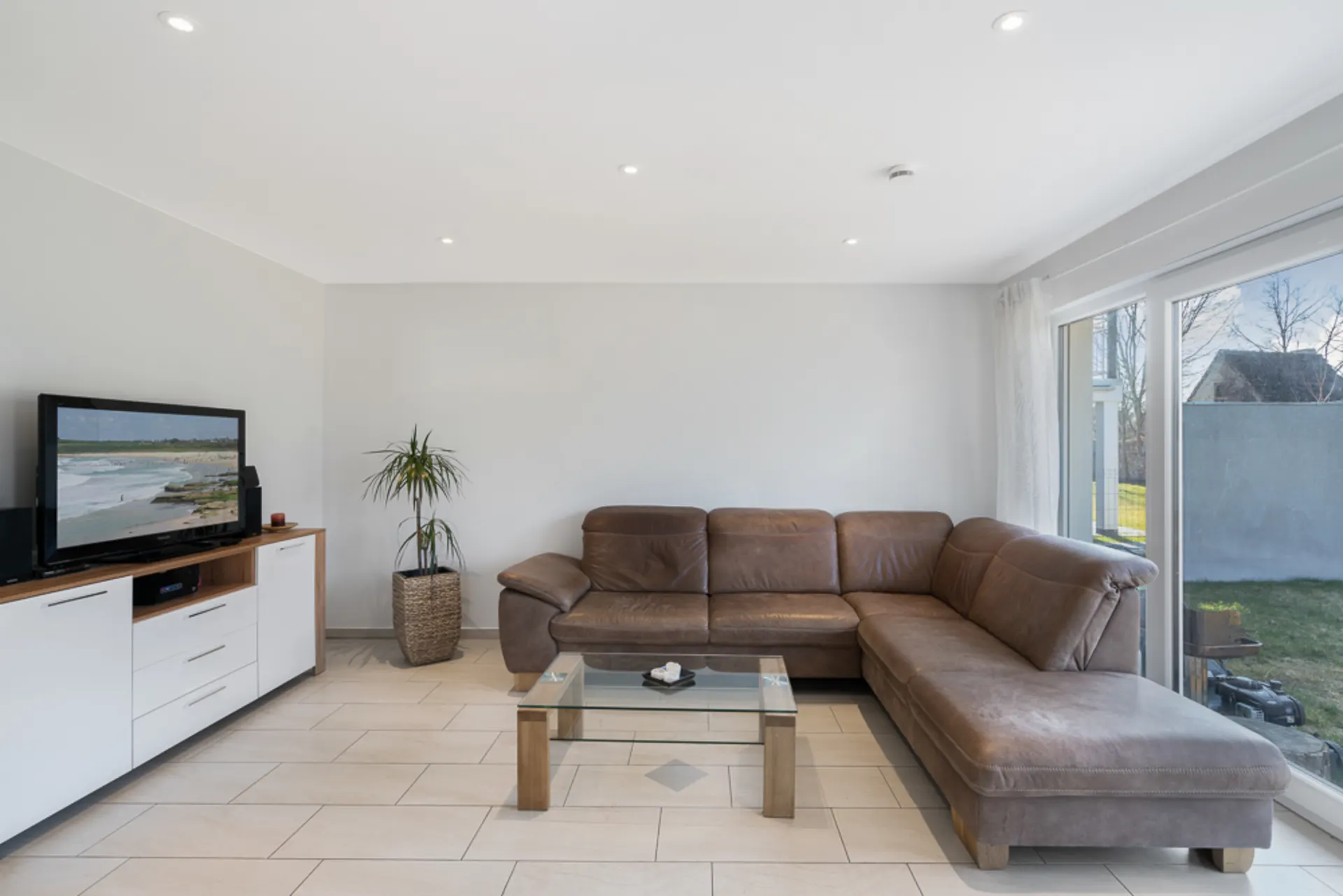 Wohnzimmer mit braunem Ecksofa, Glastisch, Fernseher und großer Fensterfront zum Garten.