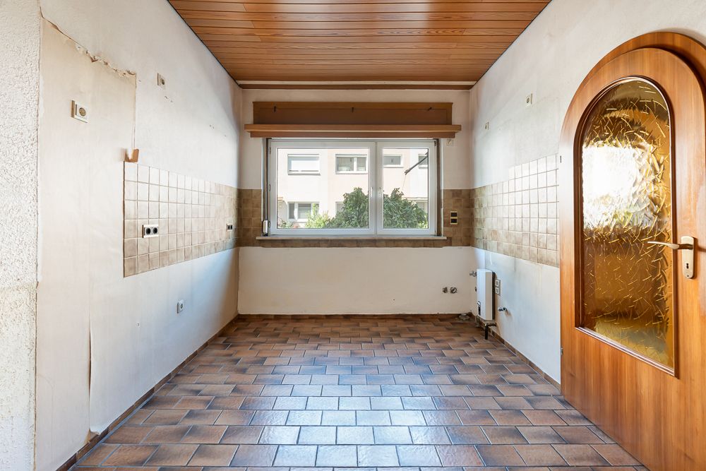Leerer Raum mit gefliestem Boden, großem Fenster und Holztür, teilweise geflieste Wände.