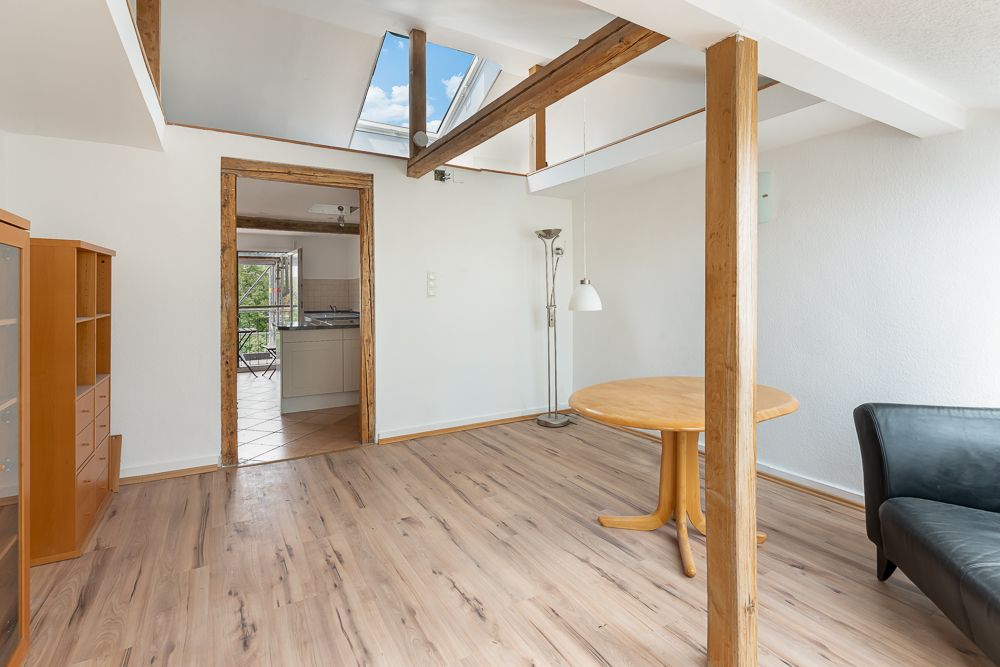 Helles Wohnzimmer mit Holzboden, Balken, rundem Tisch, schwarzem Sofa und Durchgang zur Küche.
