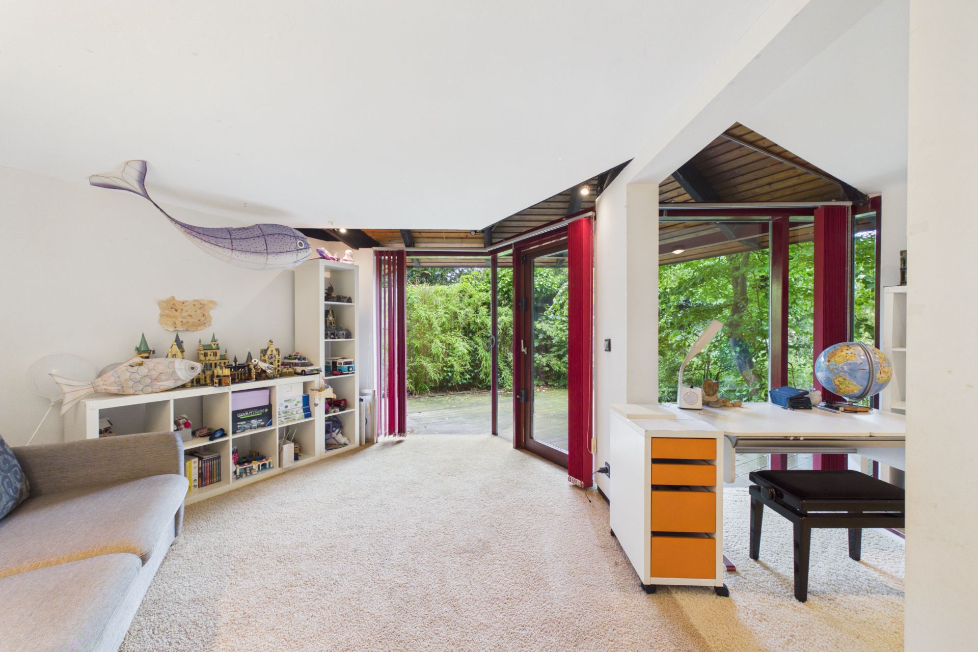 Helles Zimmer mit Schreibtisch, Regal, Sofa und großen Glasfenstern zum Garten.