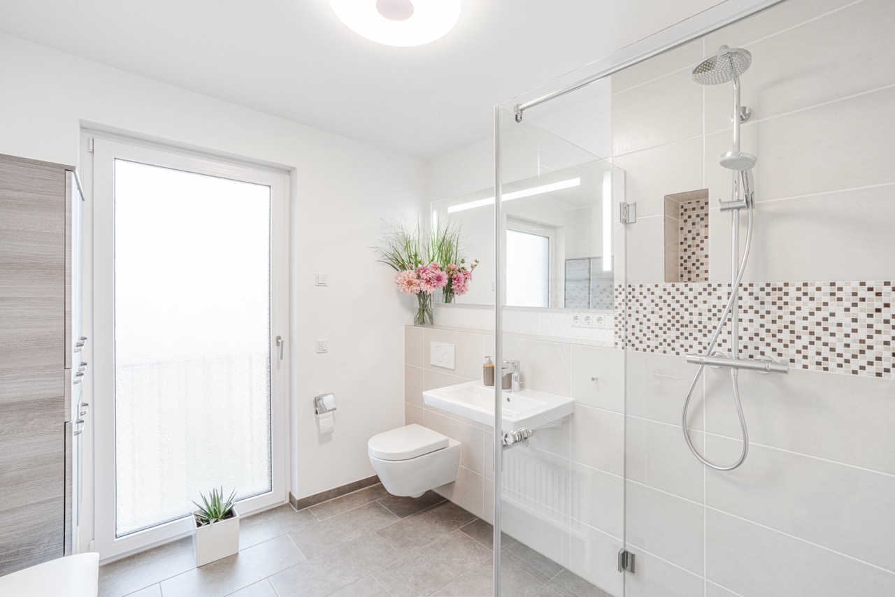 Modernes, helles Badezimmer mit Dusche, WC, Waschbecken, Blumenstrauß und Glastür.