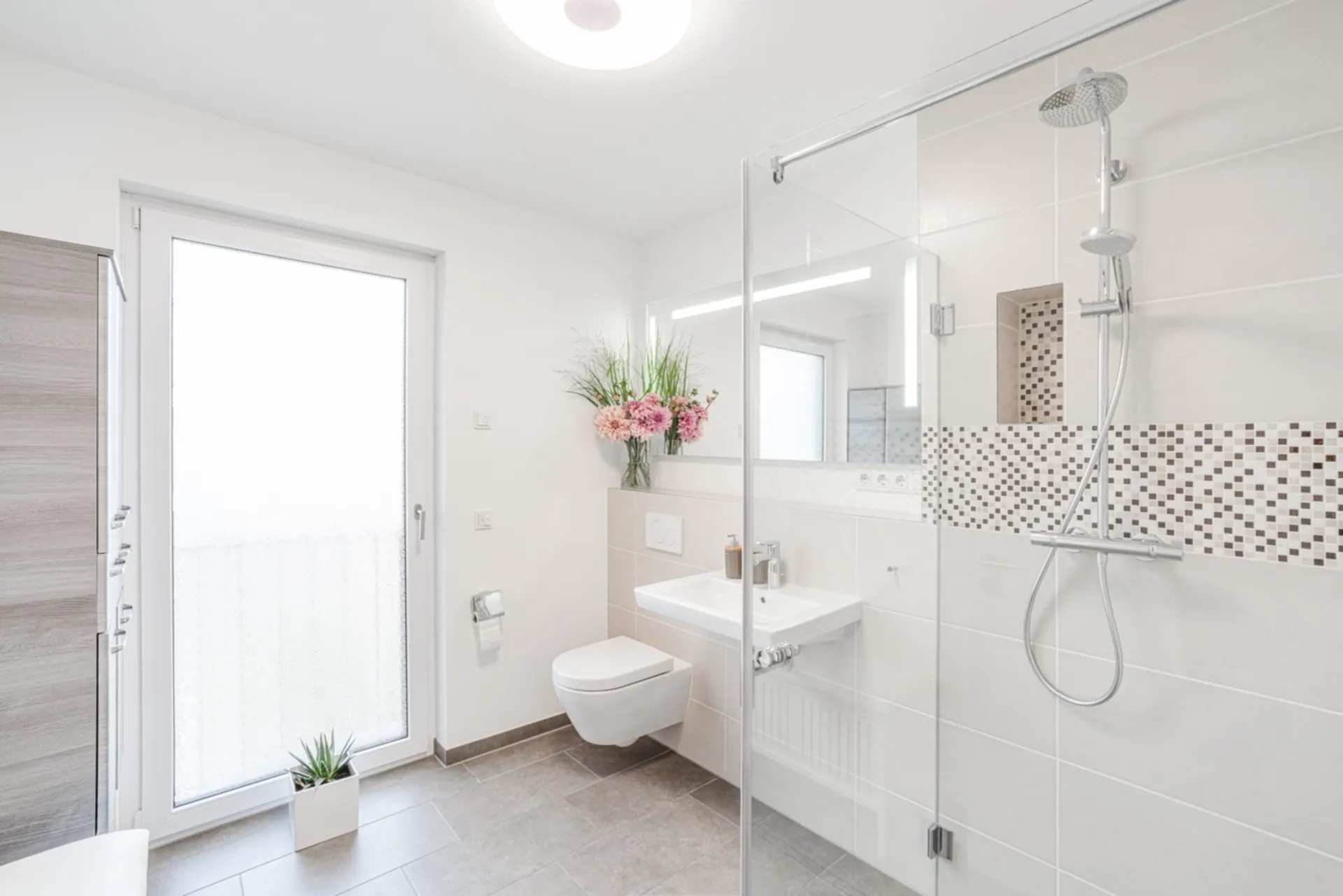 Modernes, helles Badezimmer mit Dusche, WC, Waschbecken, Blumenstrauß und Glastür.