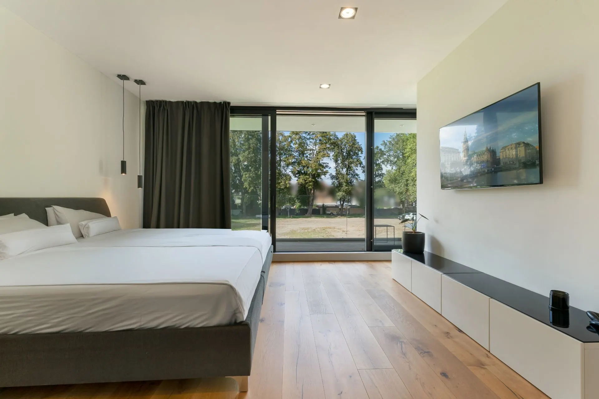 Modernes Schlafzimmer mit großem Bett, Holzfußboden und bodentiefen Fenstern mit Blick ins Grüne.