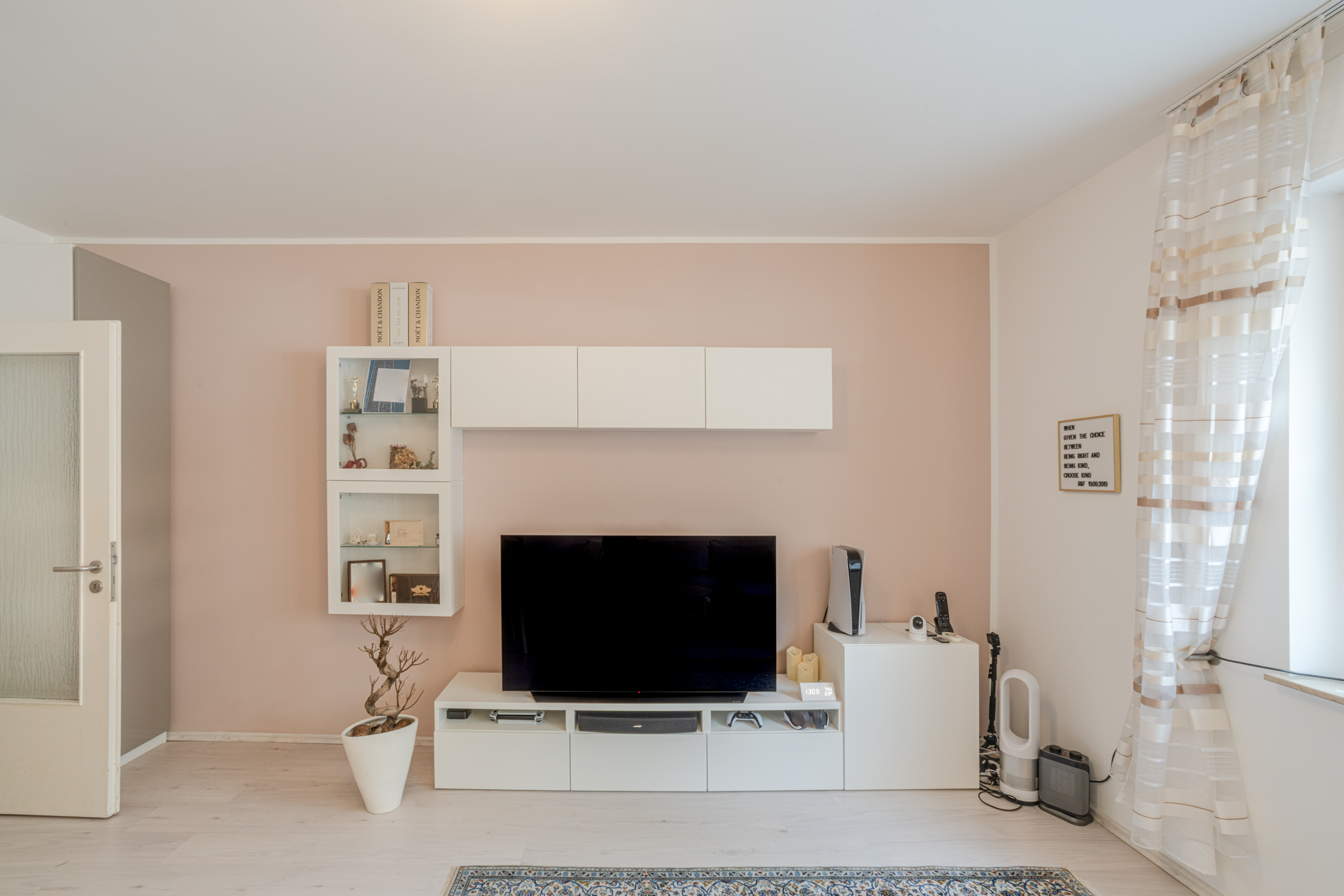 Modernes Wohnzimmer mit TV, weißen Möbeln, Regal und Pflanze vor rosa Wand.