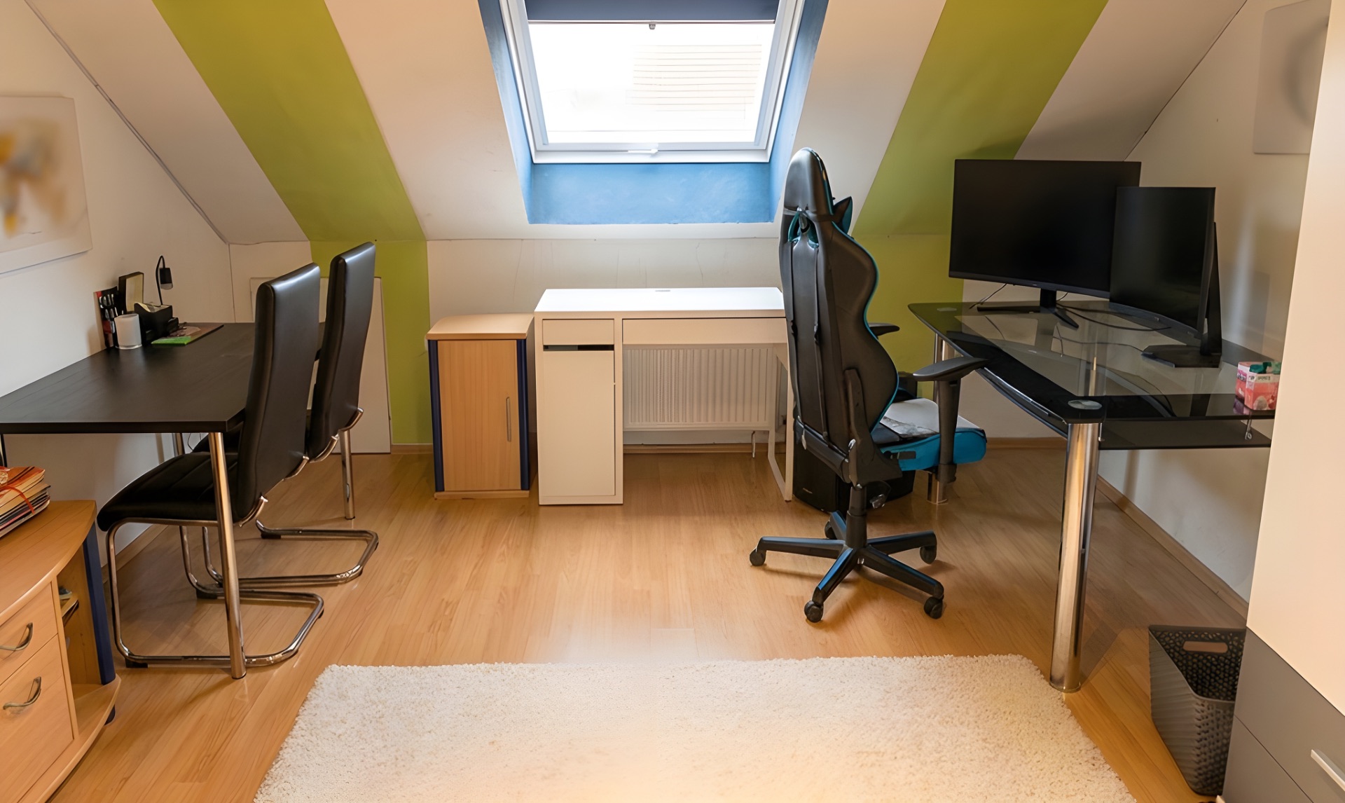 Arbeitszimmer mit zwei Schreibtischen, Bürostühlen und Monitoren unter einem Dachfenster.